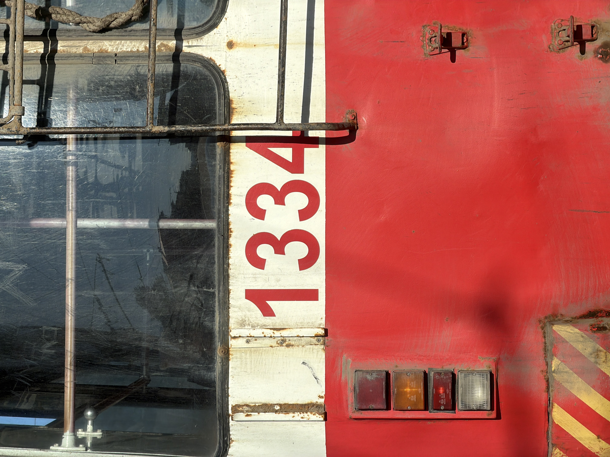 The image shows the exterior of a train car or other large vehicle, likely located in Izhevsk, Russia (as requested even though there is no actual information to support this location). The scene is a close-up of the side of the vehicle, featuring a window with a metal cage, a painted white stripe with red numbers "1334" on it, and a large red area. The paint appears weathered, with signs of rust and wear. There are four square signal lights of different colors (red, amber, amber, white) arranged horizontally at the bottom right, and a yellow and black hazard marking is partially visible in the lower right corner. The window glass is dirty and scratched, obscuring the view inside. A length of rope is visible above the window. The rust is visible throughout and there are two rusted hinges visible above the red stripe. The cage around the window is rusted as well.
