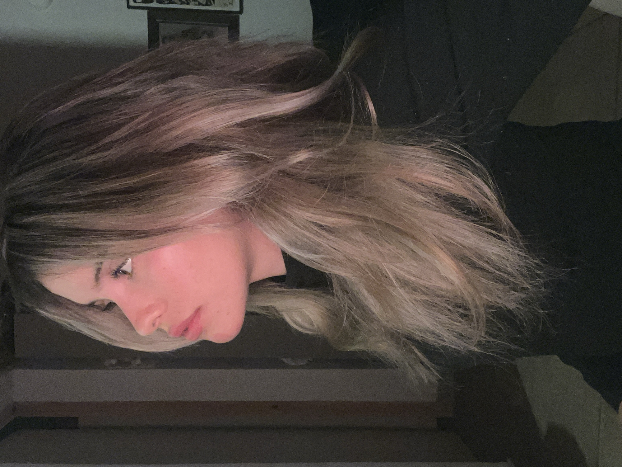 A young woman with long, wavy, blonde and brown ombre hair is shown in profile from the shoulders up, facing left. She has light skin with a pinkish hue on her cheeks and nose, and her lips are slightly parted. Her eyes are open, and she is looking upwards. She is wearing a black top. The background is dimly lit and appears to be indoors, with a doorway or frame visible on the left and a dark wall with a framed picture on the right. There are no visible elements suggesting the location is the city of Sumalao, Argentina.