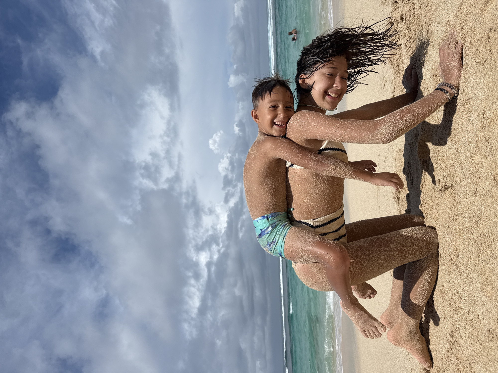 This image captures two children, a girl and a younger boy, engaged in playful activity on a sandy beach. The photo is taken from a low angle, almost horizontally, showcasing a dynamic perspective.
The main subjects are the two children, both of whom are heavily covered in light-colored sand, indicating recent play. The older child, a girl wearing a striped two-piece swimsuit, has wet, dark hair that appears windswept. She is smiling broadly and looking directly at the camera, with her left arm extended and hand pressed into the sand. The younger boy is piggybacking on her, wrapped around her back, and is also smiling widely at the camera. He is wearing patterned swim trunks.
The setting is a tropical beach, with fine sand stretching across the foreground and a calm, turquoise ocean in the background, meeting the shore with gentle waves. The sky above is partly cloudy, with patches of blue visible through the white and grey clouds. A distant figure of a person can be faintly seen in the water. The time of day appears to be daytime, likely sunny, given the bright illumination and distinct shadows. The overall emotion conveyed by both children is one of joy and happiness, reflecting carefree play. There is no visible text in the image. The location context of Kuta Selatan, Indonesia, aligns with the tropical beach scene.