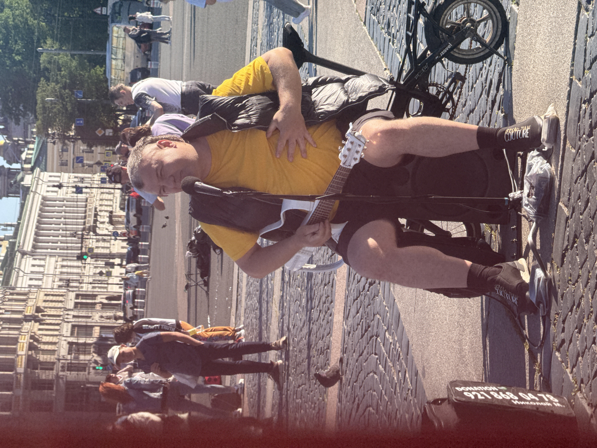 A man is seated on a chair, playing a white guitar and singing into a microphone. He is wearing a yellow t-shirt and a dark vest. The man is on a cobbled street in Saint Petersburg, Russia, with a large building in the background. There are other people walking in the background and a bicycle is attached to his chair.