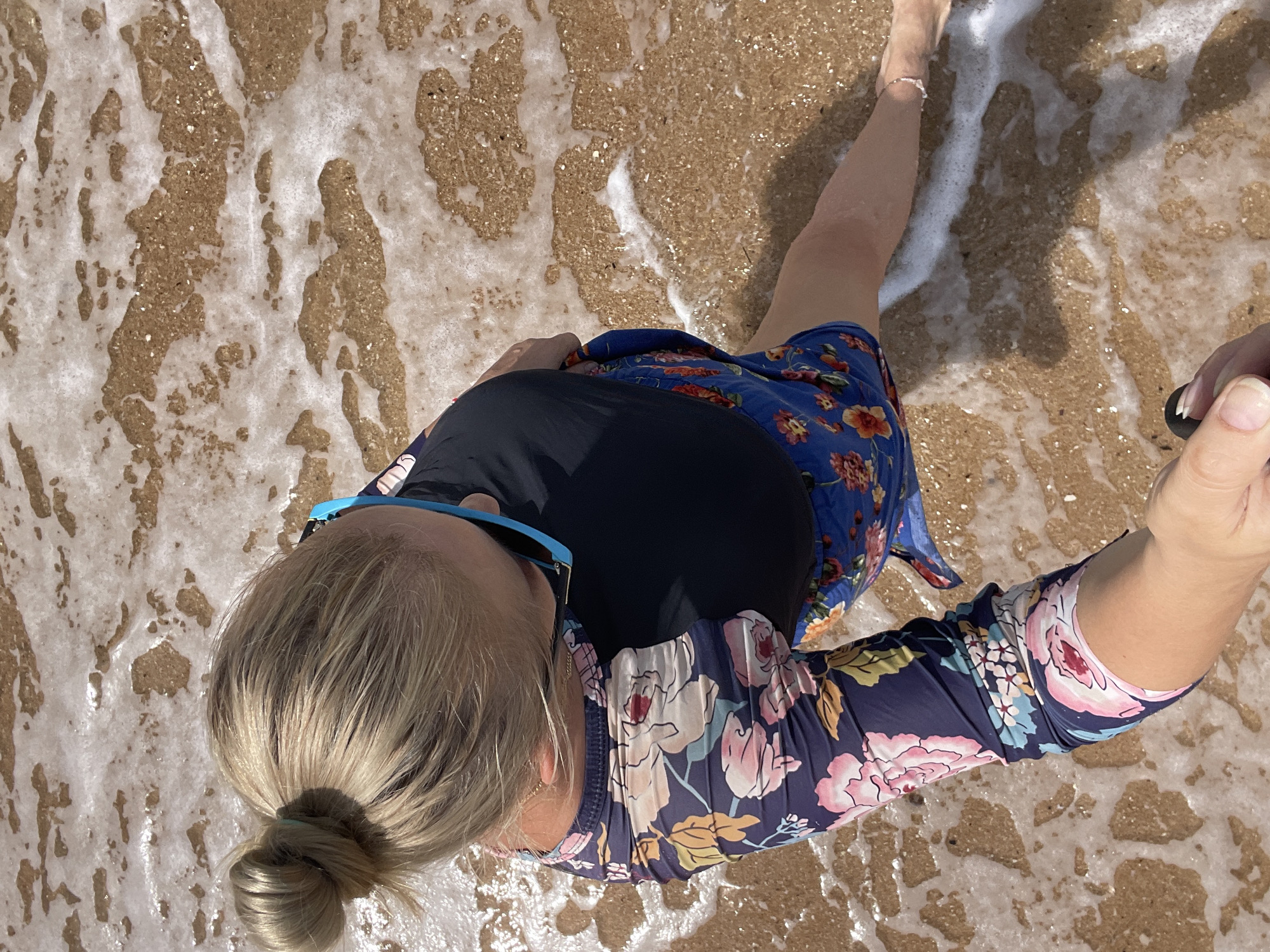 A woman, viewed from above, stands at the edge of the sea in Pattaya City, Thailand. She has blonde hair pulled up in a bun and wears blue-framed sunglasses, a black rash guard, and a blue floral patterned skirt. Her left arm, also patterned, is extended towards the viewer, holding a black selfie stick. The water laps around her feet, which are adorned with a delicate anklet. The sand beneath the water is a light brown color, and the seafoam creates a frothy white pattern on its surface. The overall scene suggests a casual day at the beach.