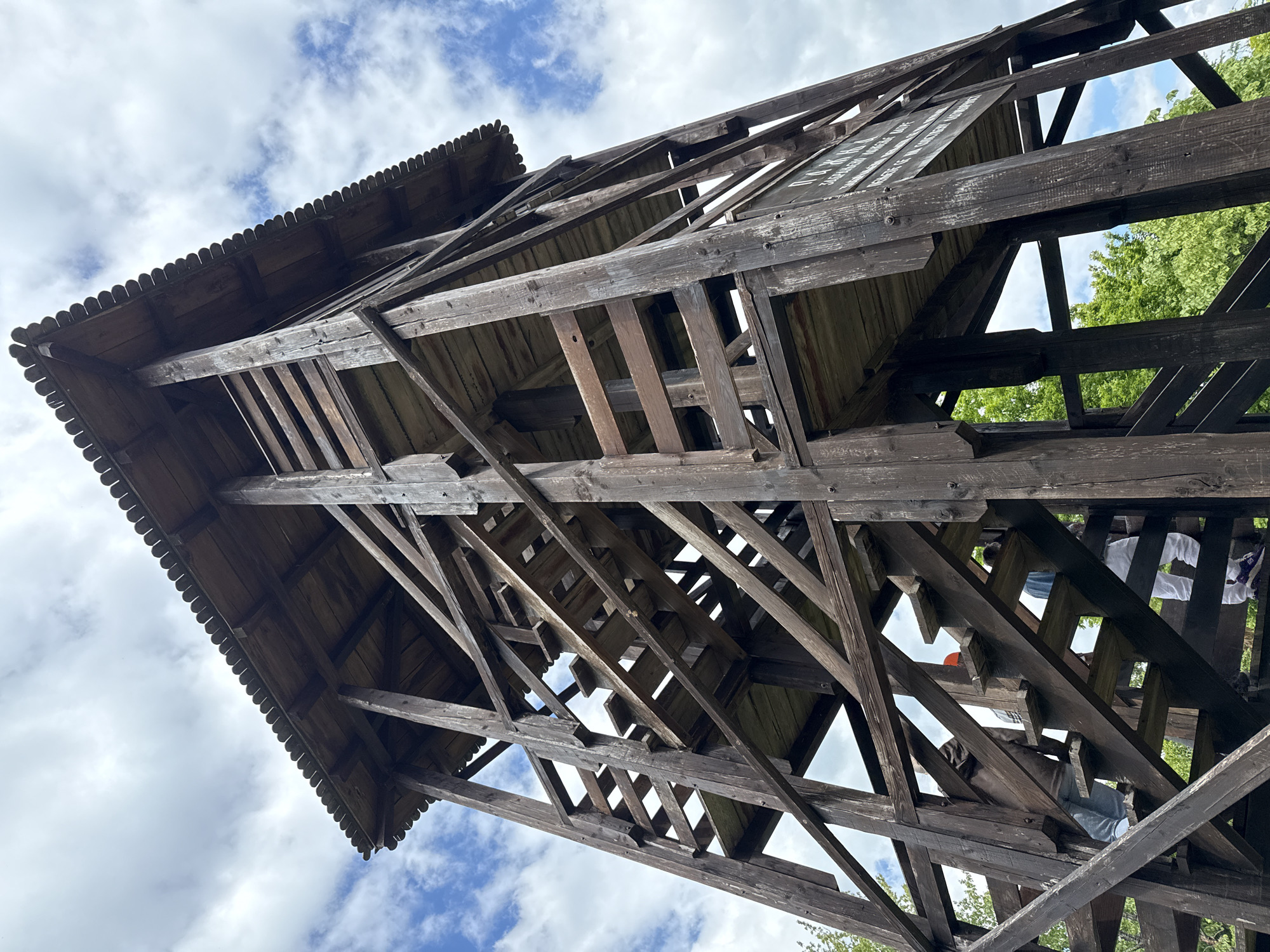 The image shows a low-angle view of a wooden structure, specifically a lookout tower. The structure is brown, and its construction is visible. Some elements are dark due to weathering.
The scene is located in Sremski Karlovci Municipality, Serbia. The image captures the perspective from below, looking up at the lookout tower, partially obscuring the sky. Patches of sky with white clouds are visible through the open framework of the structure. The top of the tower features a decorative roof edge. The tower is built on a steep angle. The stairs up the tower are visible. People are on the stairs.