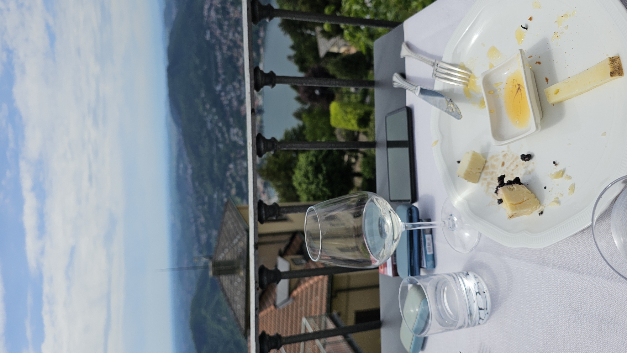 On a table in Brunate, Italy, next to a black wrought iron railing, a plate of partially consumed cheese and honey sits next to a wine glass. Below, a tile roof is visible. In the distance, a hazy blue and green landscape stretches out.