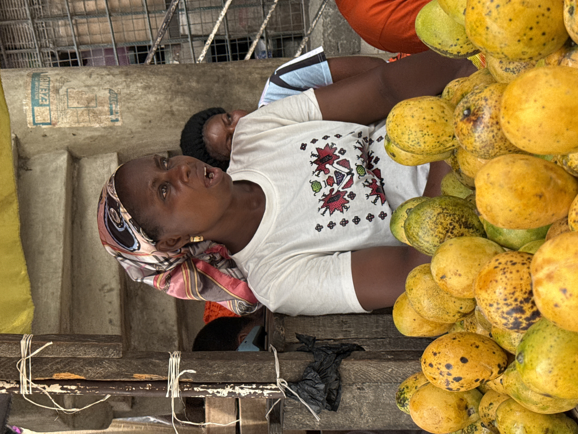 This image captures an outdoor market scene, likely in Lagos, Nigeria, featuring a woman and a large display of mangoes. The central subject is a dark-skinned woman, viewed from the chest up, looking slightly upwards and towards the viewer with an open-mouthed expression, as if speaking. She wears a colorful patterned head wrap, a gold earring, and a white t-shirt adorned with red, black, and green embroidered motifs.
To her right, partially obscured, a child wearing a dark cap is visible, appearing to be asleep or resting. In front of the woman, a substantial pile of mangoes is displayed. These mangoes vary in color from bright yellow to green, many with dark speckles, suggesting different stages of ripeness. They are stacked high, indicating a plentiful supply.
The setting includes weathered wooden planks tied with white string in the immediate foreground, forming part of a stall or display. Behind the woman is a light-colored concrete wall, on which a faded light blue rectangular label or sticker is visible. Part of the text on this label is discernible, showing "OMEZ" and other less clear letters. In the background, beyond the wall, metal grating or fencing suggests an urban environment. The scene is brightly lit, indicating daytime. The overall impression is one of daily commerce in a bustling, outdoor setting.