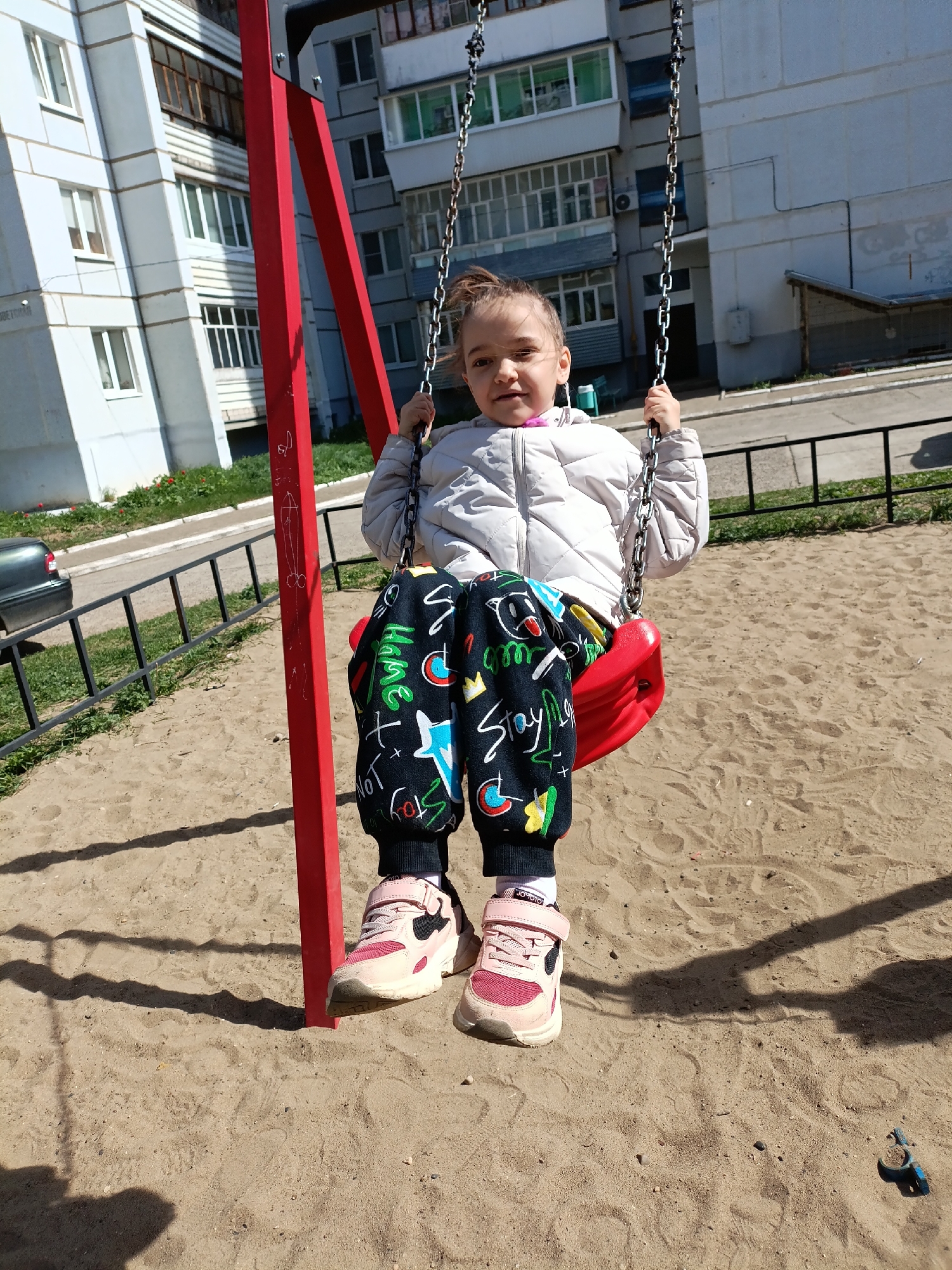 A young girl is sitting on a red swing in a playground. The girl is wearing a light-colored quilted jacket and black pants with colorful designs. She has pink shoes. The playground is covered in sand. The swing set has a red metal frame. In the background are apartment buildings, a metal fence, and a parked car. The location is the city of Chaikovsky, Russia.