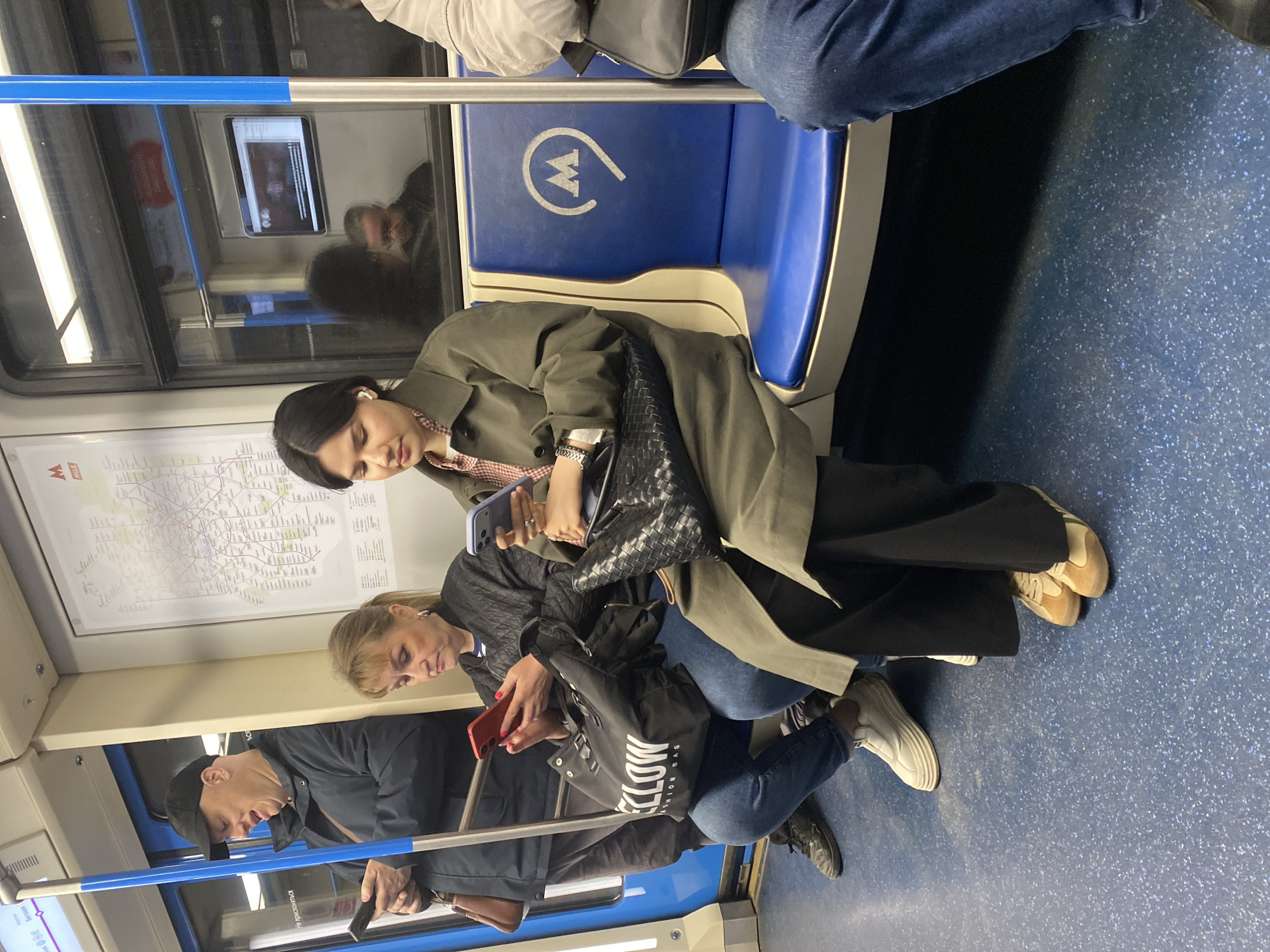 The image depicts a scene inside a subway car, likely in Khimki, Russia, based on the prominent "M" logo on the blue seat, indicating the Moscow Metro system. Several people are visible. A young woman with dark hair is sitting, wearing a green coat and holding a phone. An older woman next to her is also using a red phone. A man wearing a black cap is standing, also holding a phone. The subway car has blue speckled flooring, blue seats with a white "M" logo, and a map on the wall. People are focused on their phones, suggesting a common activity during commutes.