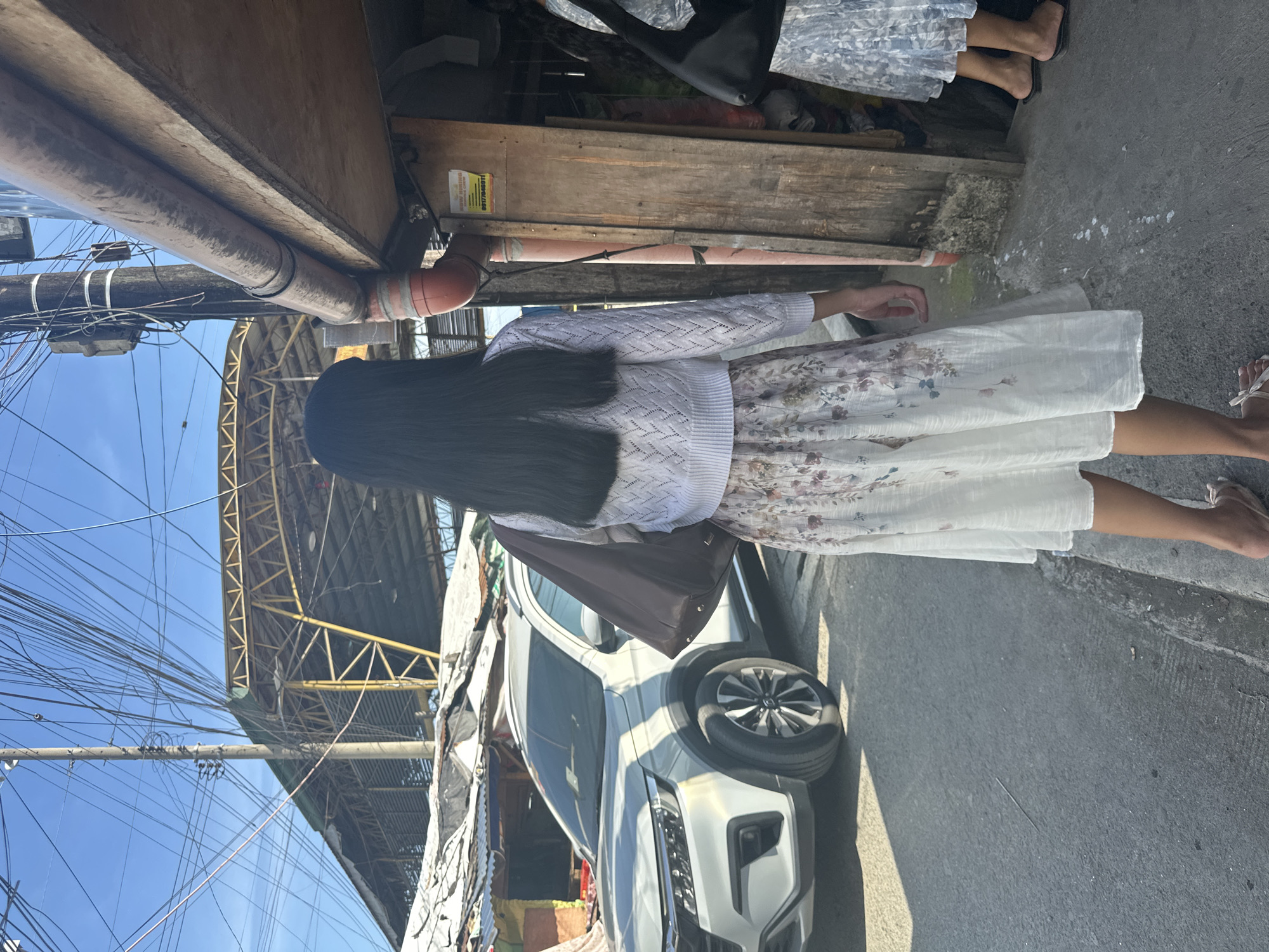 A person with long, dark hair, seen from behind, is walking away from the camera on a street. They are wearing a white knit cropped cardigan over a white skirt with a floral pattern. A brown shoulder bag is slung over their left shoulder. To the left of the person, a white SUV is parked on the street. In the background, there are market stalls with tin roofs and a large, yellow, covered structure. The scene is outdoors, with a clear blue sky visible. The overall environment suggests an urban setting, possibly a market area, in Davao City, Philippines, indicated by the presence of the SUV and the style of the market stalls.