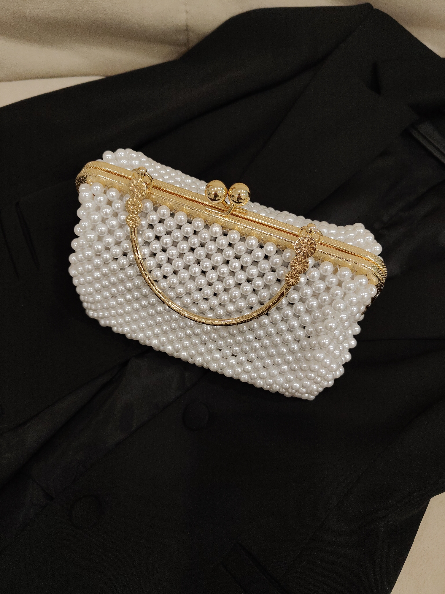 A pearl-covered purse with a gold frame and handle rests on a black blazer. The scene is indoors, location unknown.