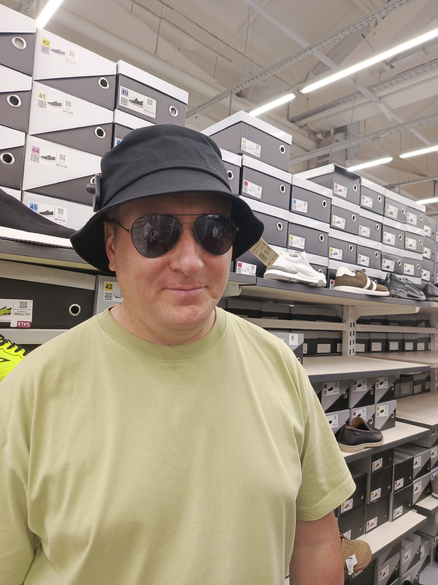 In Chaikovsky, Russia, a man wearing a black bucket hat, sunglasses, and a light green t-shirt stands in an athletic shoe store. He faces the camera. Behind him are shelves stacked with athletic shoe boxes and displayed shoes. The shoe boxes are primarily dark gray, with white labels indicating sizes and models. Shoes in various colors and styles are on the shelves. Overhead, the store is illuminated by fluorescent lighting.
