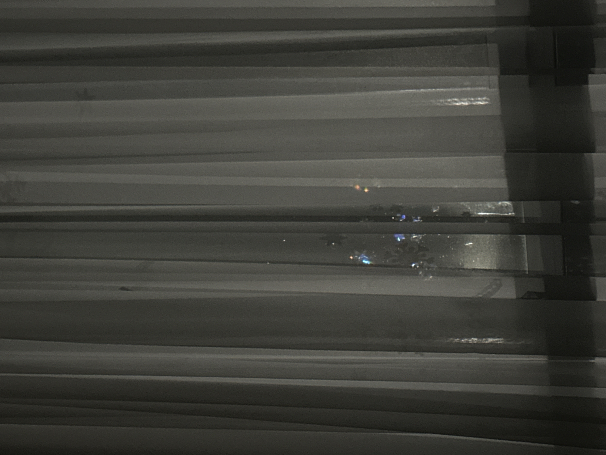 The image depicts a close-up view through window blinds, revealing a blurry cityscape at night. The blinds are horizontal and mostly closed, with a few slats slightly open, allowing glimpses of lights from distant buildings. The scene is dimly lit and details outside the window are unclear due to the focus on the blinds and the darkness. There are no people visible in the image. The location is unknown.