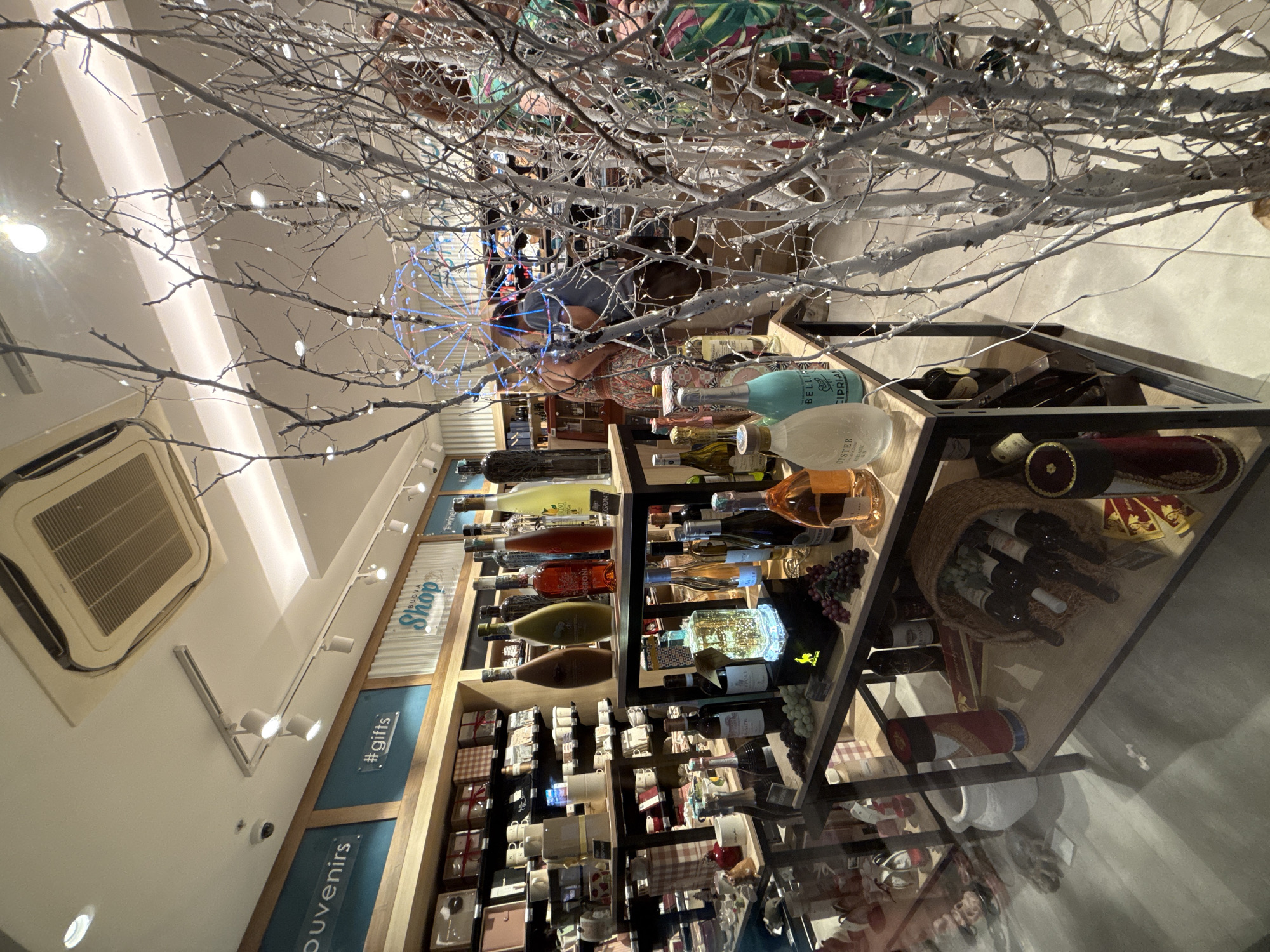 The image depicts the interior of a shop in Budva, Montenegro, likely a gift shop or liquor store, with a focus on alcoholic beverages and souvenirs. The scene is characterized by shelves stocked with various bottles of wine and spirits, gift boxes, and decorative items. In the foreground, a display of wines is prominently featured, with some bottles illuminated by internal lights. To the right, a decorative tree with white branches and small lights adds to the ambiance. In the background, people are visible browsing the merchandise. The shop appears well-lit, with overhead lighting and spotlights illuminating the displays. Signs indicating "Souvenirs," "#gifts," and "Shop" are visible on the walls, suggesting the nature of the establishment. The overall impression is of a commercial space designed to attract shoppers with a variety of products and a pleasant atmosphere.