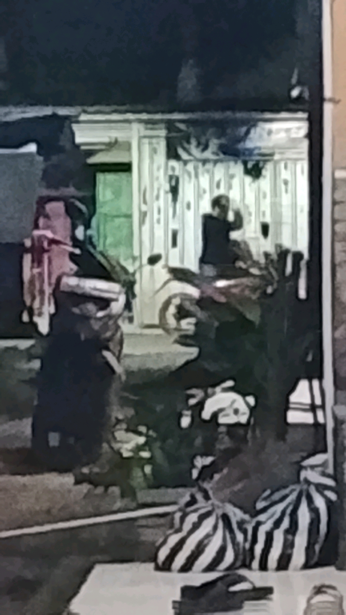 The image shows a man in a dark shirt standing near a motorcycle in what appears to be an outdoor area. The background consists of a building with striped siding and a green door. In the foreground, two large striped bags are placed on the ground, and a pair of flip-flops is visible. The scene is dimly lit, suggesting it may be nighttime or an indoor space with poor lighting. The overall impression is of a residential or commercial area in Ujungrusi, Indonesia.