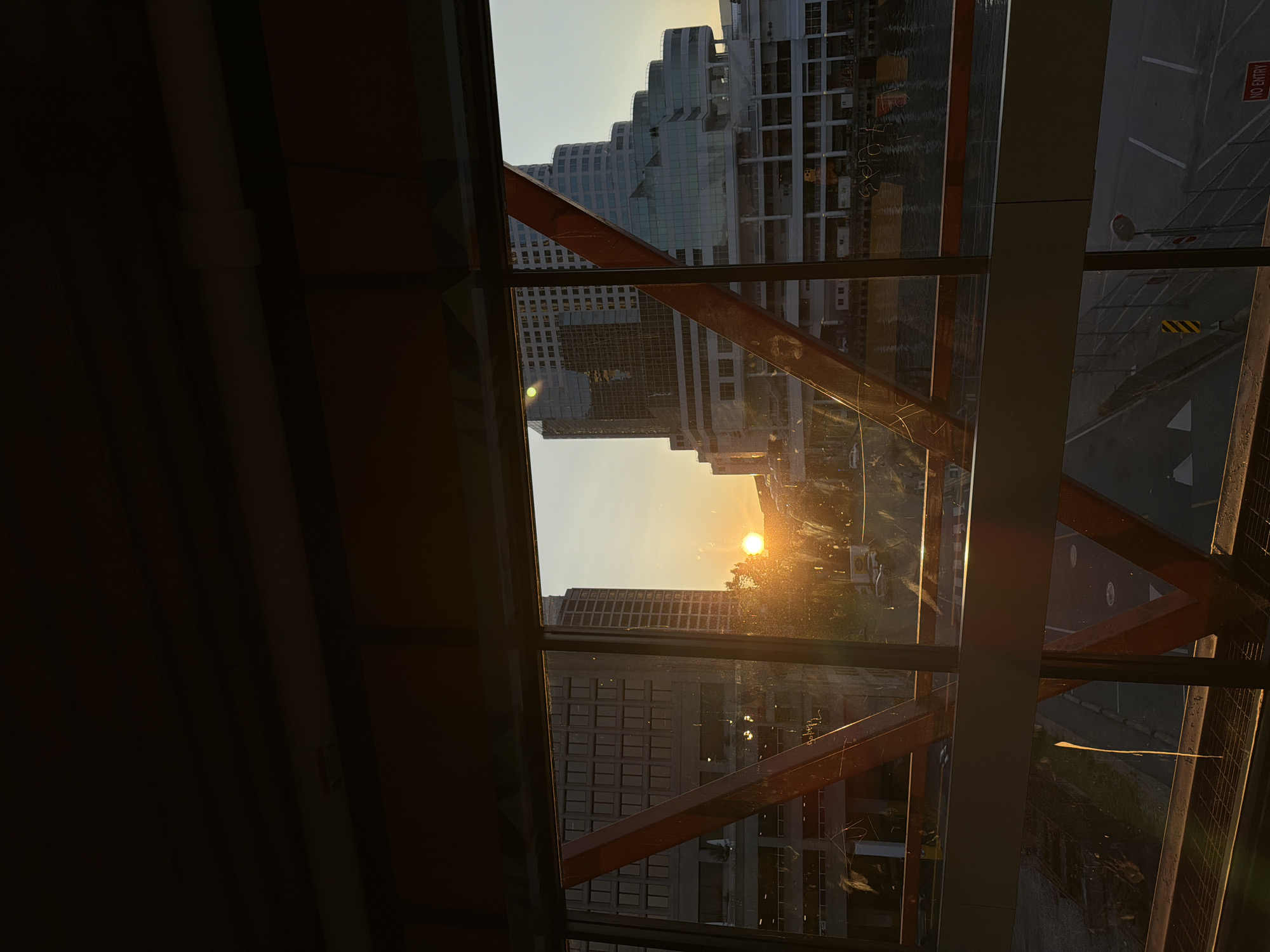 The image captures a sunset viewed through the glass and metal structure of a skywalk in Vancouver, Canada. The sun, a bright orange orb, is low in the sky, casting a warm glow and lens flares across the scene. Two modern high-rise buildings dominate the background. The building on the left is a rectangular, dark-glass structure, while the building on the right is a more complex, tiered design with lighter glass facades. Below the buildings, a body of water, likely a harbor or inlet, is visible, with some vessels or structures in the water. The foreground features the orange-colored metal framework of the skywalk, with glass panels reflecting the sunlight and the surrounding environment. The sky is clear and shows a gradient of warm tones. The overall impression is one of an urban sunset, seen from an elevated vantage point.