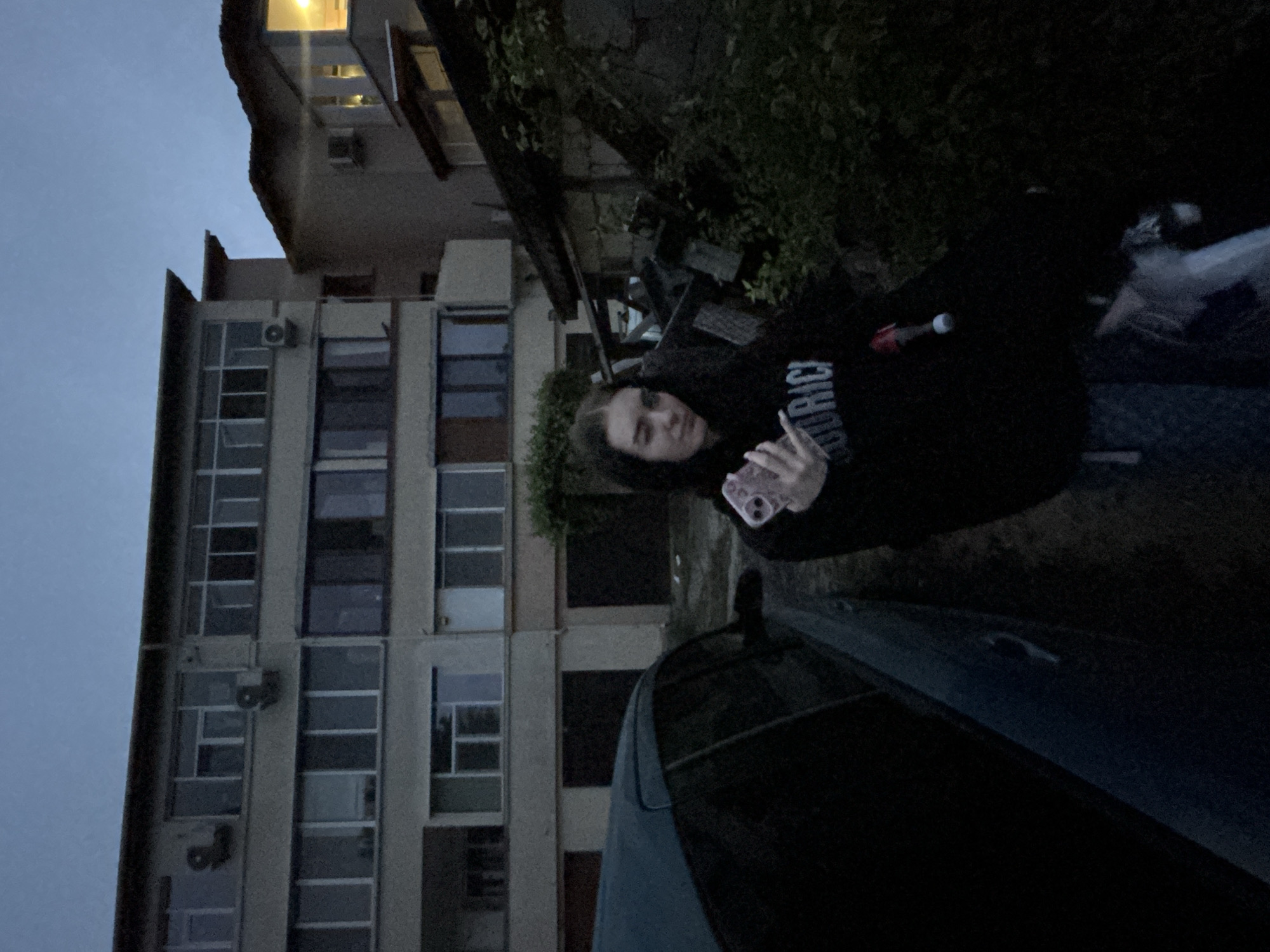 A medium shot, taken outdoors at dusk, shows a woman standing near a dark car in the city of Burgas, Bulgaria.
The woman is facing the camera, holding a smartphone. She is wearing a dark hoodie with visible writing on the front.
The backdrop features a multi-story building with a grid of windows, some of which have air conditioning units attached. Another building is visible in the upper portion of the frame. Lush green vegetation obscures part of the buildings. The sky is overcast.