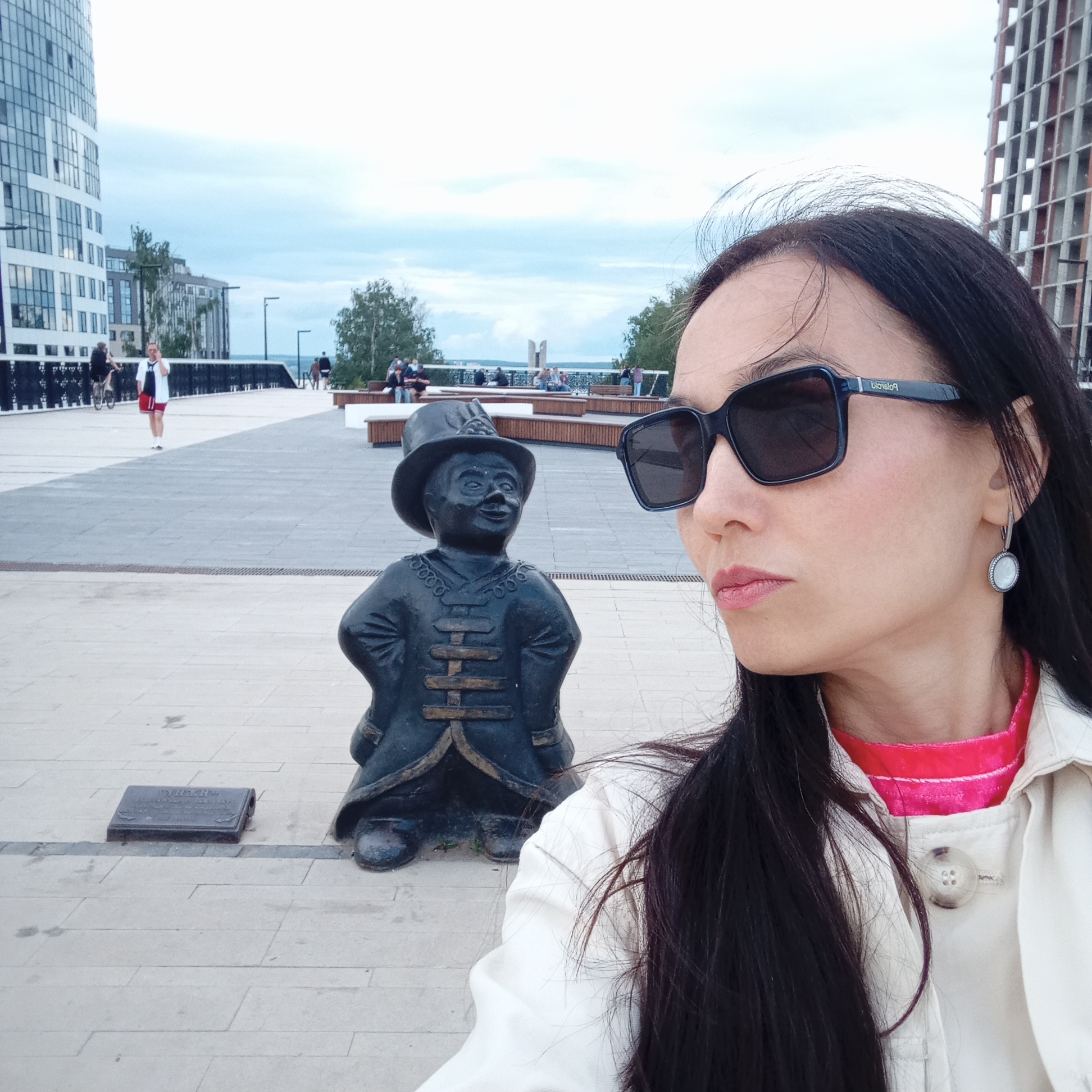 The image captures a selfie featuring a woman in the foreground and a statue in the midground, set in an urban outdoor environment identified as the Izhevsk Pond Embankment in Izhevsk, Russia.
In the immediate foreground, a woman with long, dark hair is visible from the right side, facing left. She wears large, dark rectangular sunglasses with "Polaroid" visible on the right temple arm. Her lips are painted a light red or pink, and she wears a white buttoned jacket over a bright pink top. A silver-colored earring with a circular white or light-colored stone is visible on her right earlobe. Her expression is neutral to slightly serious. She appears to be holding a camera or smartphone to take the picture, with her right arm possibly extended.
To the left of the woman, centrally positioned in the midground, is a dark, possibly bronze or cast iron, statue known as "Izhik." The statue depicts a small boy or man wearing a hat, a stylized coat with horizontal fastenings, and boots. He stands with his hands on his hips and a wide, smiling expression on his face. At the base of the statue, on the light grey paved ground, lies a small, rectangular, dark metal plaque.
The background reveals a wide pedestrian promenade paved with light and dark grey tiles. To the far left, a tall, modern building with a facade of numerous windows is visible. Further back, on the right, another building with a grid-like structure under construction is partially visible. In the middle distance, several individuals are present; one person in a white top and red shorts is walking towards the foreground, while another on a bicycle is moving away. Farther back, people are seen sitting on benches and walking along the embankment. A body of water, likely the Izhevsk Pond, is visible under a cloudy sky in the distance. Green trees are interspersed along the embankment, and a distinctive monument with two upward-curving elements is discernible in the far background, overlooking the water. Streetlights are placed along the promenade.