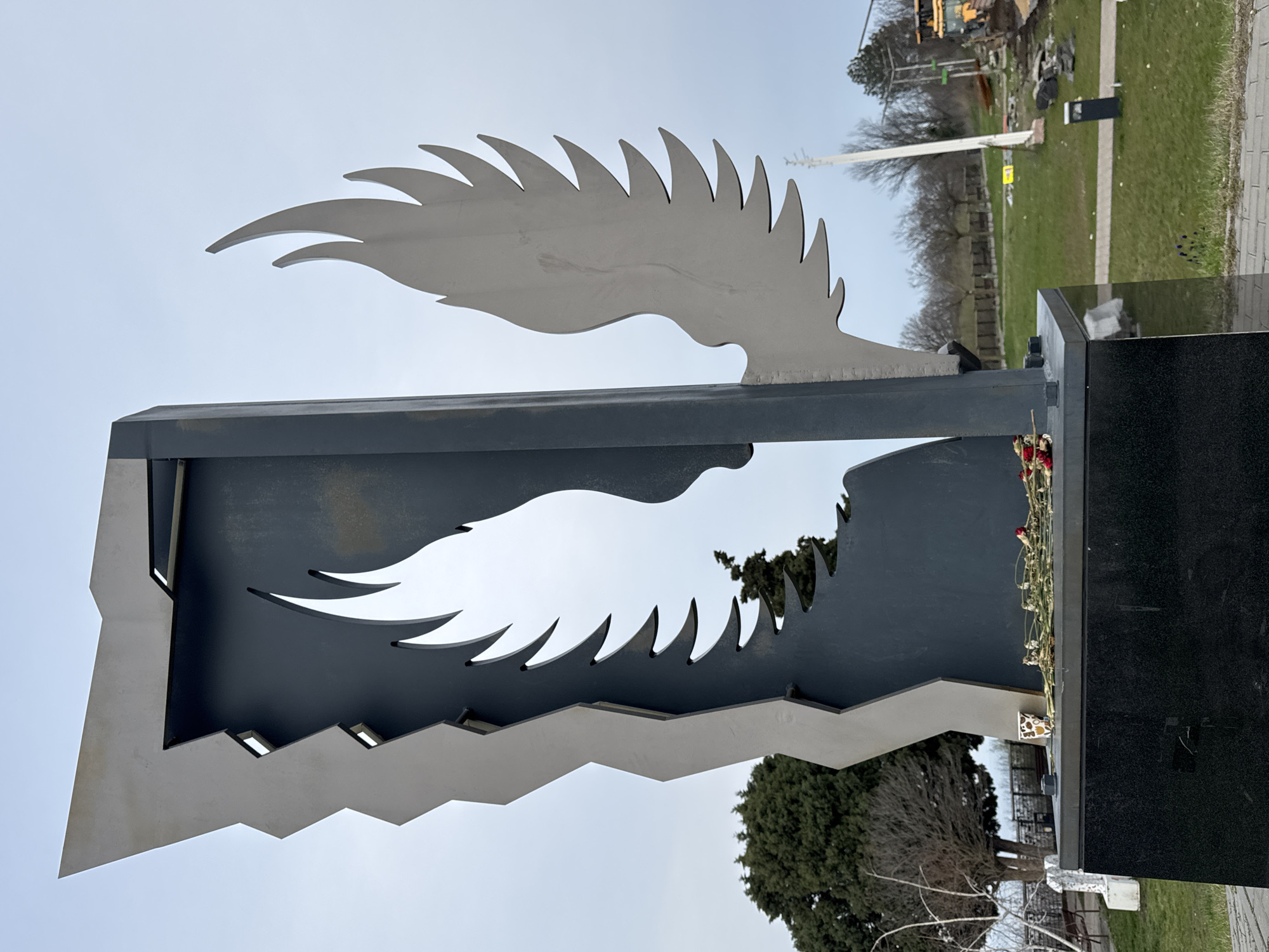A low-angle, close-up shot of a black and silver memorial in a grassy area. The memorial has a stylized metal wing extending upwards and another cut-out wing shape within a dark grey rectangular metal structure. To the right of the dark grey structure is a polished black pillar. There are dead flowers laid against the pillar. The sky is overcast. In the background, there is green grass, trees and construction equipment. This memorial is located in Varna, Bulgaria.