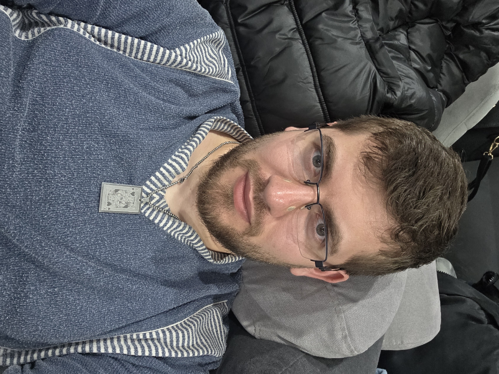 A man with a beard and glasses lies on a grey pillow against a black quilted jacket. He wears a blue sweater with white stripes at the collar and shoulders, and a silver necklace with a card-shaped pendant. The location is unknown; Yagodovo, Bulgaria cannot be verified from the image.