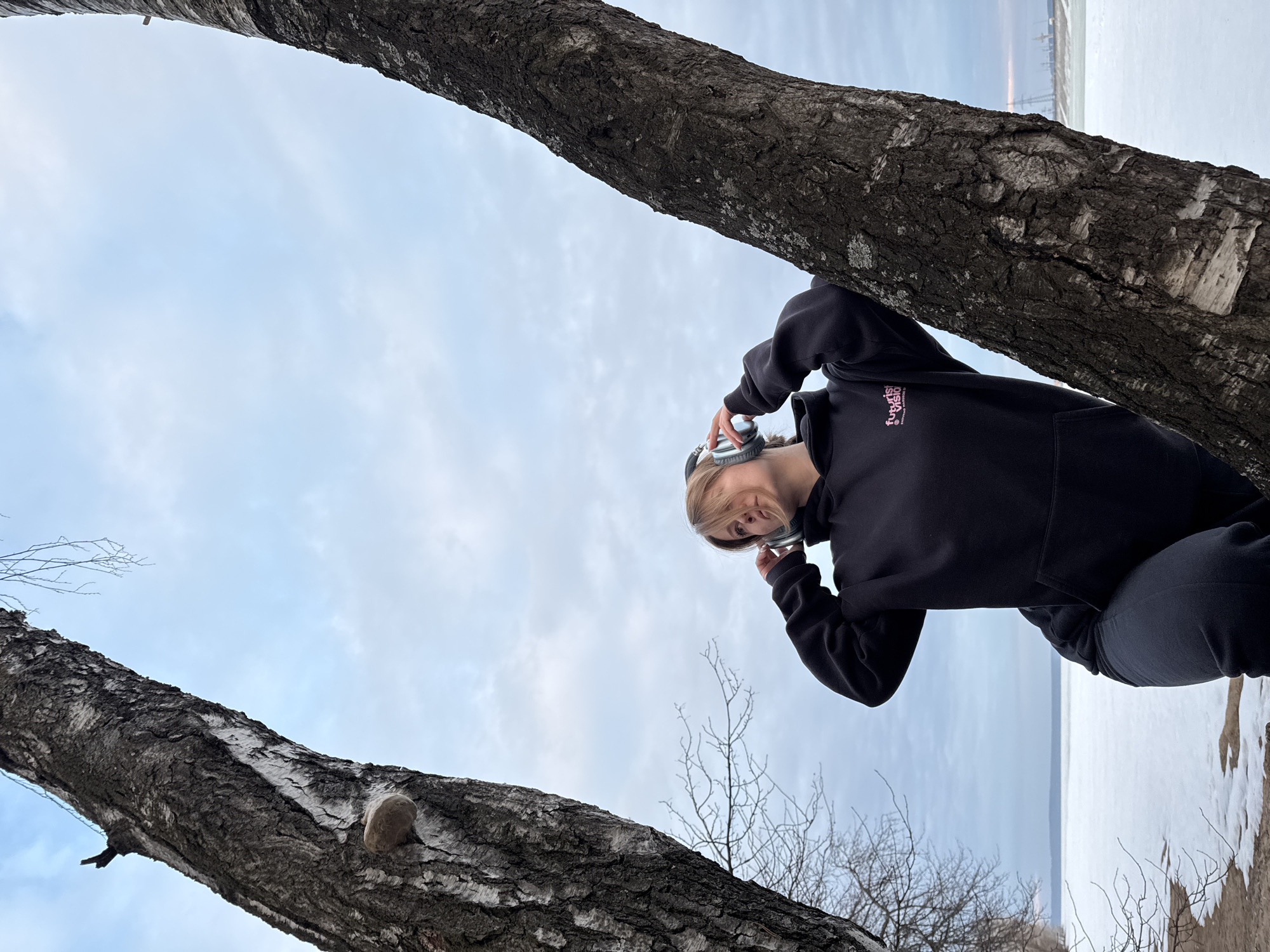 The image displays a lone individual in Noviy, Russia, positioned outdoors between two prominent, dark-barked tree trunks, possibly birch. The subject, a female with light-colored hair, is wearing a black hooded sweatshirt with a pink and white logo on the left chest, and dark trousers. Both hands are raised, holding silver over-ear headphones over her ears, with her gaze directed towards the upper right. A fungal growth is visible on the left tree trunk. The immediate foreground features a narrow strip of dark earth, transitioning to a vast, flat expanse of snow and ice covering a body of water in the middle-ground. Bare tree branches are visible against an overcast, light blue-grey sky. In the far distance across the frozen water, on the right horizon, indistinct industrial structures are visible. The scene is set in a cold environment, indicative of winter or early spring conditions.