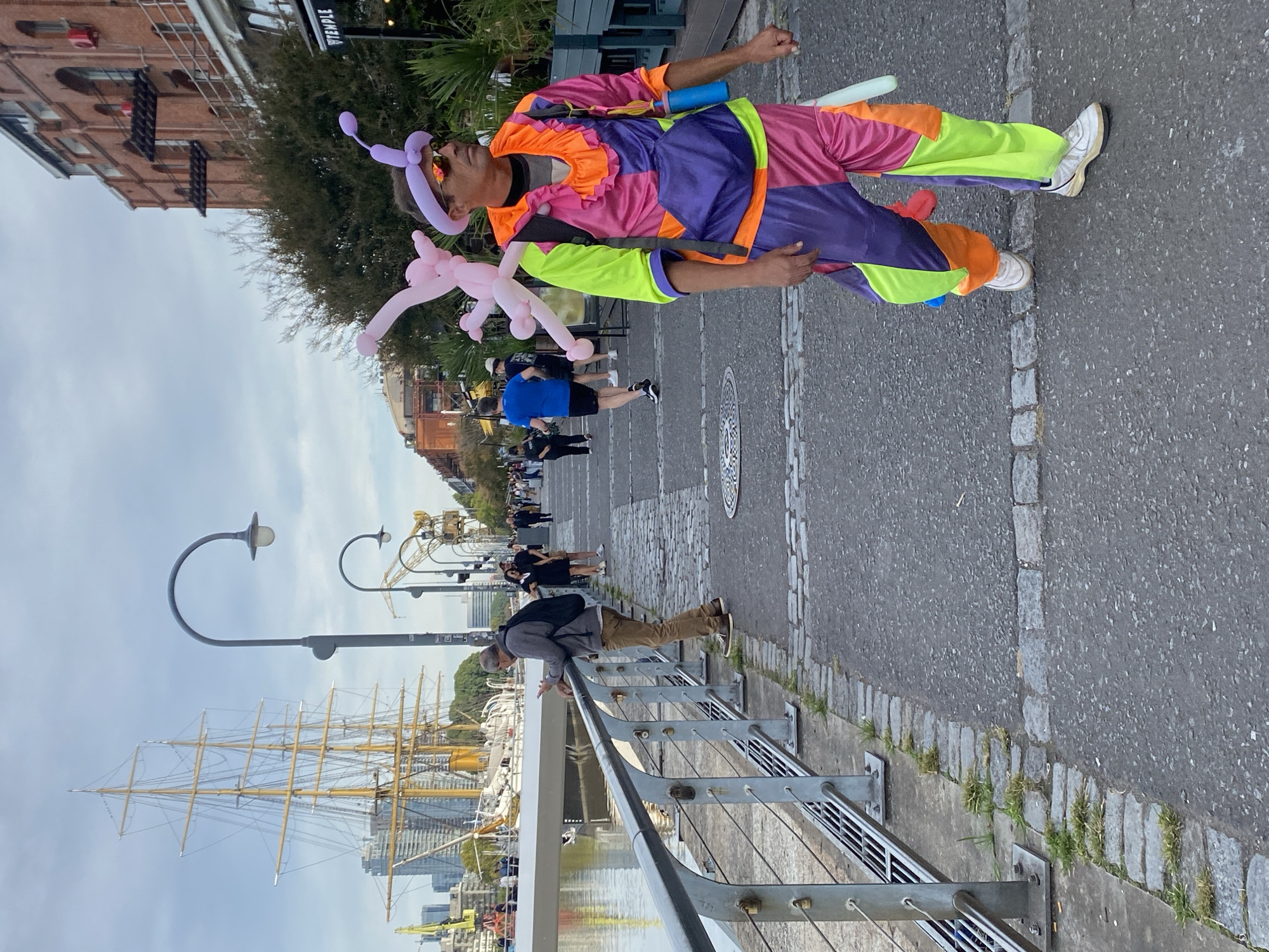 A man dressed in a colorful clown suit, with a balloon animal on his head and a pink balloon animal in his hand, is standing on a gray concrete sidewalk next to a metal railing. Other people are walking in the background. A large sailing ship is docked on the water behind the railing. A light pole and streetlamps are on the sidewalk next to the railing. In the background are various buildings, including a brick building with a black sign hanging from the front. The image appears to be taken in Puerto Madero, Buenos Aires, Argentina.