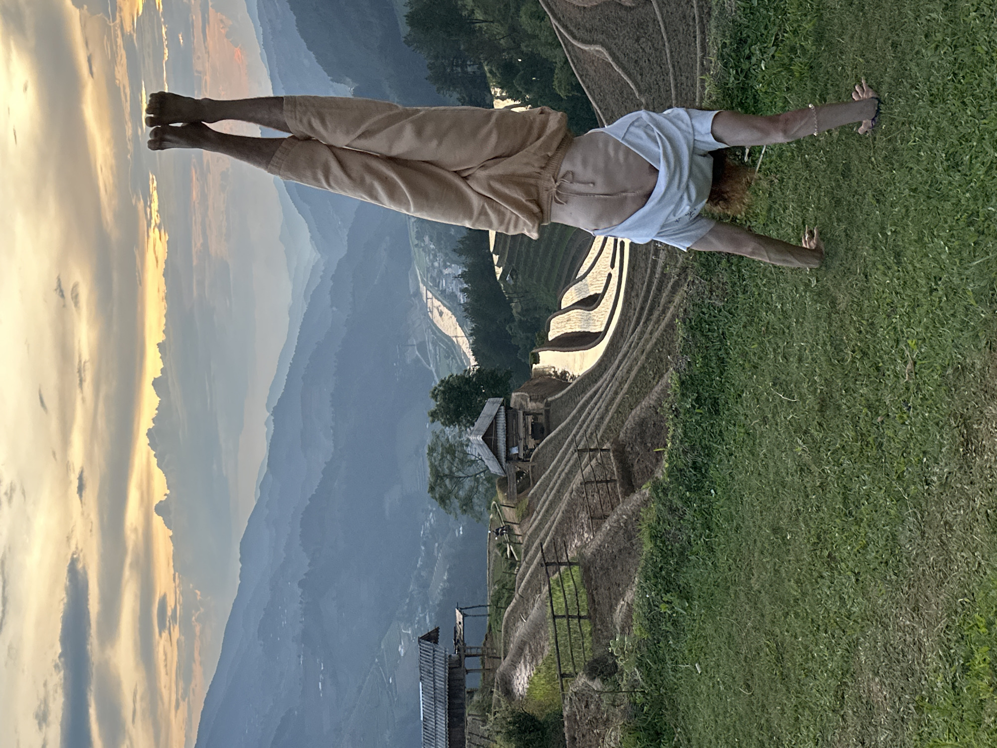 The image captures a person performing an inverted pose, likely a handstand or yoga inversion, on a grassy slope, with their back to the viewer. The individual has light brown, curly hair and is wearing a light-colored t-shirt and beige trousers. Their bare feet are pointed upwards, and their hands are firmly placed on the vibrant green grass.
The scene unfolds as a breathtaking landscape of tiered rice terraces, characteristic of mountainous agricultural regions like Mù Cang Chải, Vietnam. The intricate, undulating patterns of the terraces, some appearing wet and reflective, cascade down the mountainside. Amidst these terraces, small, traditional wooden houses with distinctive roofs are nestled, partially obscured by trees. A wide valley with a winding river or body of water stretches into the distance, framed by multiple layers of misty mountains.
The time of day is golden hour, either sunrise or sunset, casting a warm, soft glow across the sky and illuminating the scattered clouds with shades of gold and orange. The weather appears clear and calm. The image conveys a sense of peace and connection with nature through the physical activity set against such a majestic and serene backdrop. No visible text is present in the image.