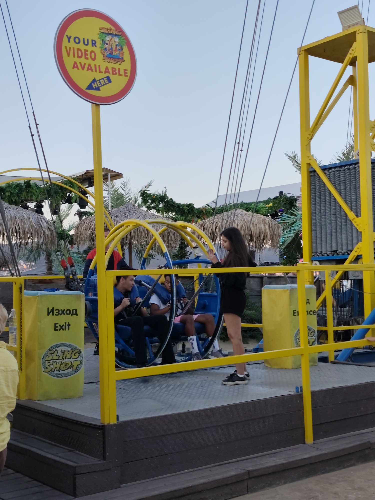 In the city of Nesebar, Bulgaria, an amusement park attraction features a bright yellow metal frame and a ride with blue seats. A circular red and yellow sign with the text "YOUR VIDEO AVAILABLE HERE" and an illustration of people on a ride is mounted on a yellow pole. Several people are visible on the attraction, some wearing blue restraints. Two large yellow pillars with the word "EXIT" and the logo "SLING SHOT" written on them are present. Another yellow pillar with the word "EN" is also visible. In the background, palm trees and thatched-roof structures suggest a tropical or resort-like atmosphere. The sky is clear and blue.