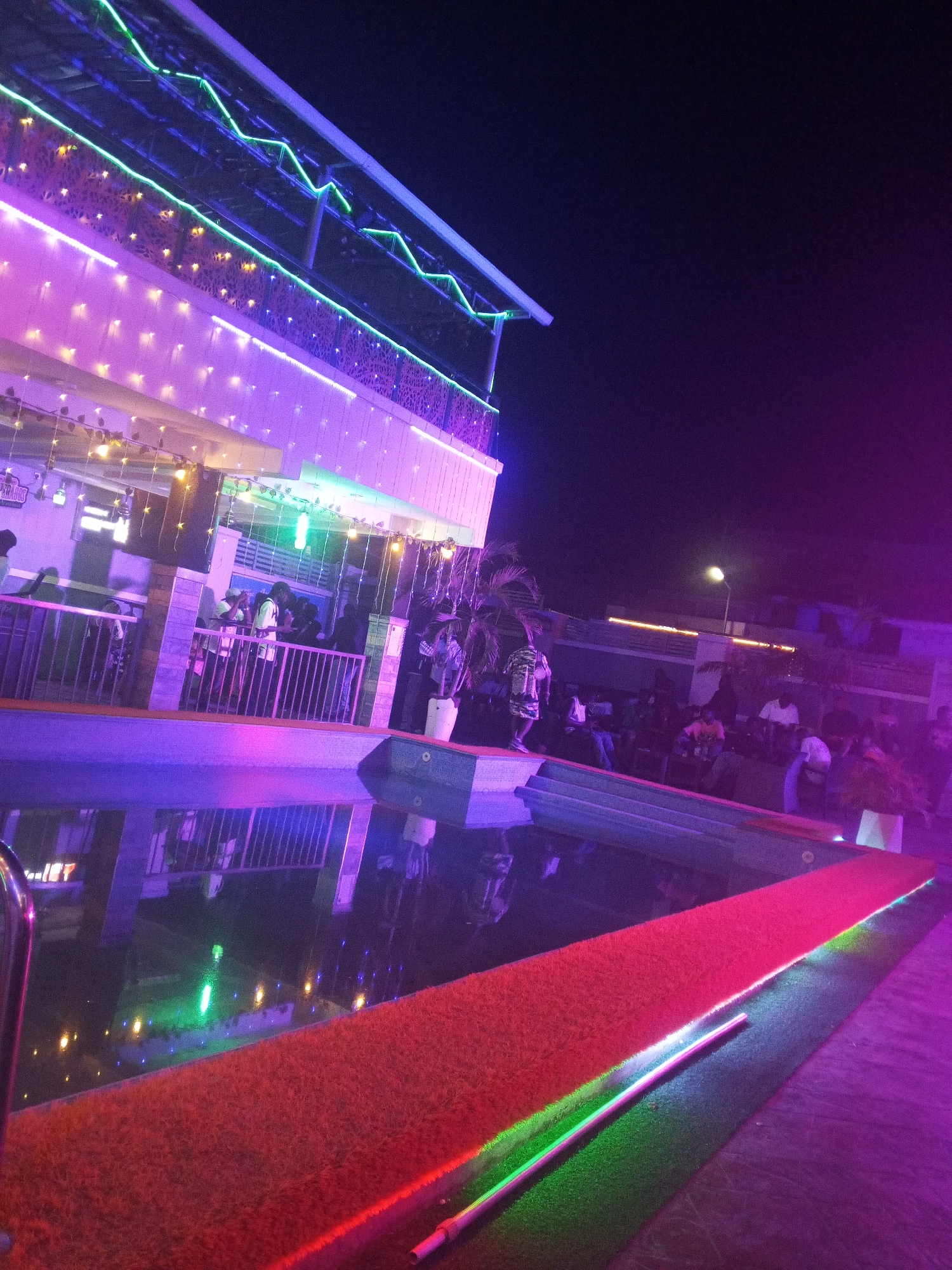 The image depicts an outdoor night scene, likely a social gathering or event. The location is possibly in Lokoja, Nigeria, though this cannot be confirmed from the visual information alone. A two-story building is visible, adorned with decorative lights along its exterior, including neon-style lights. There is a pool in the foreground, with a red carpet or covering adjacent to it. The pool reflects the lights and surroundings, creating distorted patterns. People are present on the balcony of the building and around the pool area. Some are standing, while others appear to be seated. The lighting in the scene is predominantly purple and green, contributing to a vibrant atmosphere. A streetlight can be seen in the background. The overall environment suggests a recreational or entertainment venue.