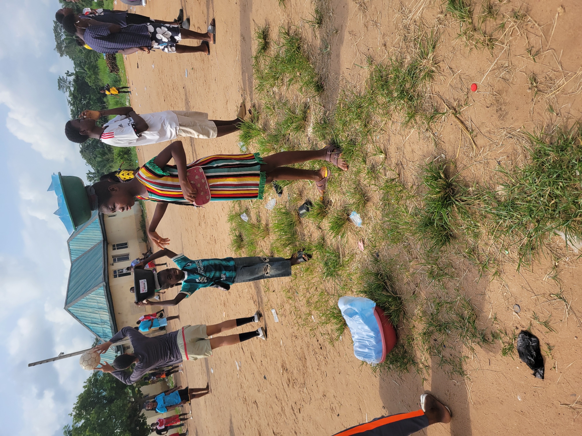 In a dusty outdoor area with sparse grass and scattered litter, a group of people are gathered. In the foreground, a young girl with a colorful striped dress balances a green bowl filled with blue objects on her head. To her left, a boy in a teal jersey is visible. Behind them, a taller person in a grey shirt and shorts holds a soccer ball above their head, facing away from the camera. Further in the background, other individuals are present, some in athletic wear. A light-colored building with a blue-tiled roof stands to the left. The overall setting appears to be a community gathering or event in a rural or semi-urban environment in Egbagu, Nigeria.