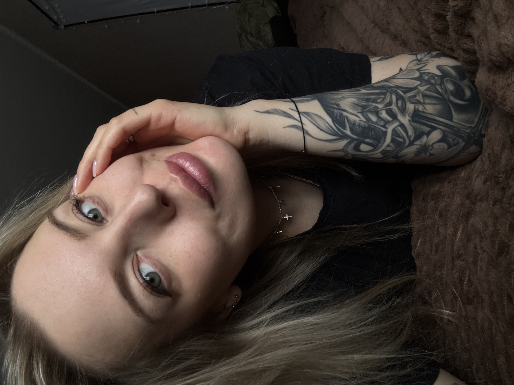 A light-skinned woman is reclining on a brown, textured surface (likely a blanket or plush fabric). She wears a black shirt. Her right arm, adorned with a large, intricate tattoo, rests on the surface beside her head, which is tilted slightly upward. Her left hand is near her face, with her fingertips touching her forehead. She has long, blonde hair. She wears a delicate necklace with small cross pendants. Her facial expression is relaxed, and she is looking directly at the camera. The location in Borzya, Russia is not apparent in the image.