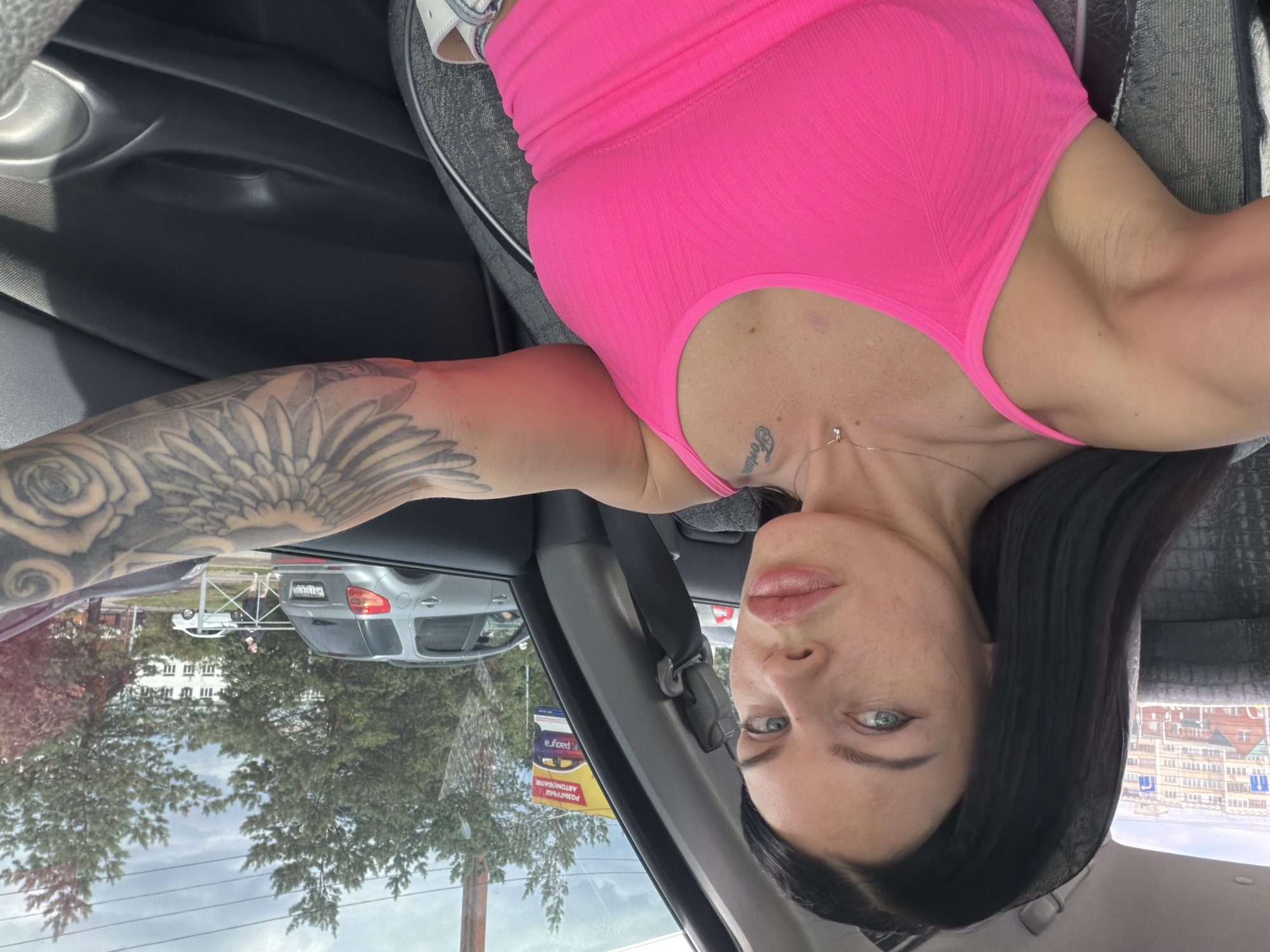 This is a selfie taken from inside a car, likely in Ufa, Russia. The image is oriented sideways, with the subject's head towards the bottom. The subject is a woman with dark hair, fair skin, and blue eyes, wearing a bright pink tank top. She has a large floral tattoo on her left arm and a small tattoo on her neck. She is looking towards the camera with a neutral expression.
The background shows the interior of the car, including a black car door and a grey car seat. Through the car window, a street scene is visible. There are trees, buildings, and other cars. One car in the foreground appears to be a silver SUV. A yellow sign with red lettering is visible on a pole, with some text that is partially obscured and appears to be in Cyrillic. The sky is overcast, suggesting it might be a cloudy day. The overall lighting suggests it is daytime. The scene appears to be a suburban or urban environment.