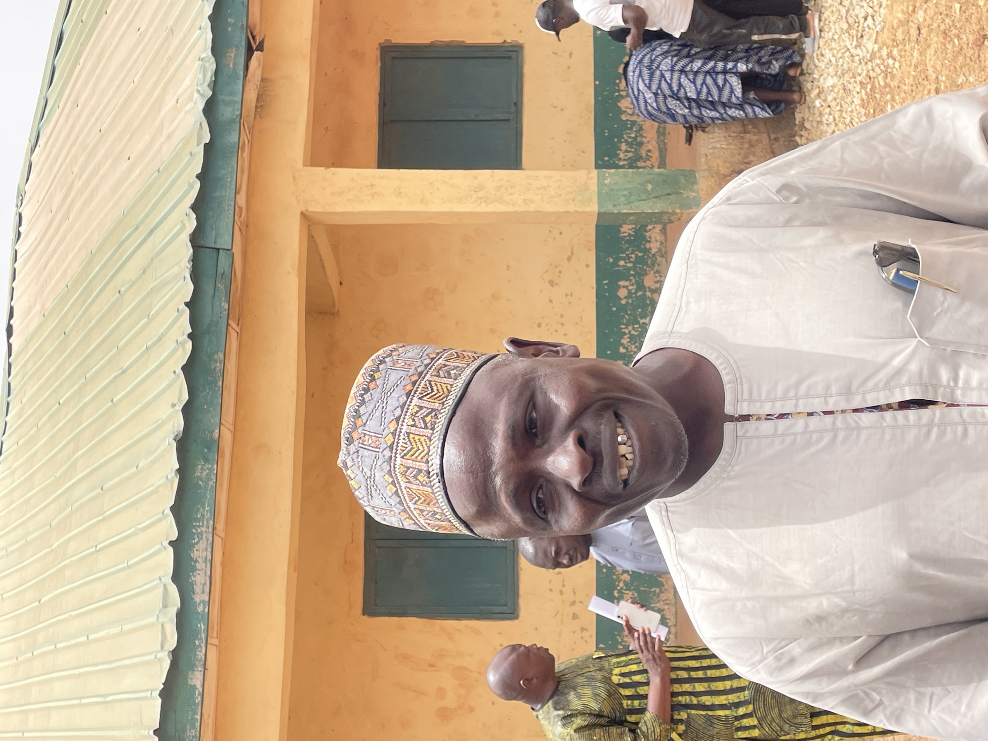 A close-up shot depicts a man wearing a patterned hat and light-colored clothing, likely in Jos, Nigeria, based on the attire and building architecture. He is positioned in front of a building with yellow walls and green window frames. Other individuals are visible in the background, along with a building with a corrugated metal roof.