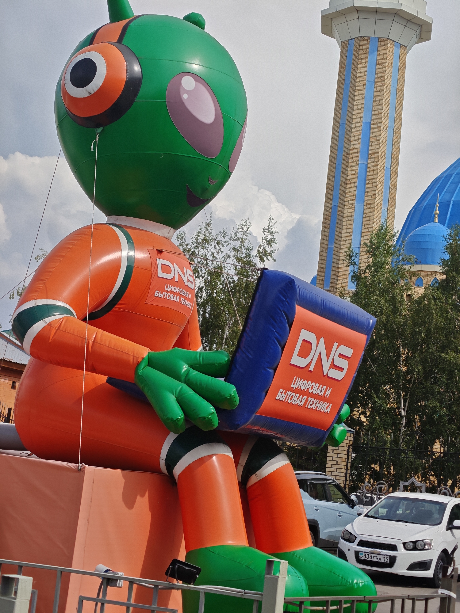 The image shows a large, inflatable green and orange alien character holding a blue laptop. The alien is positioned outdoors, with a building that appears to be a mosque, characterized by its blue dome and tall minaret with vertical blue stripes, visible in the background. The scene also includes trees, a fence, and parked cars. The license plate on the white car is visible and appears to be from Kazakhstan. The presence of the mosque and the Kazakh license plate strongly suggest the location is Petropavl, Kazakhstan. The alien figure is advertising for "DNS," which is a Russian electronics retailer. The sky is partially cloudy.
