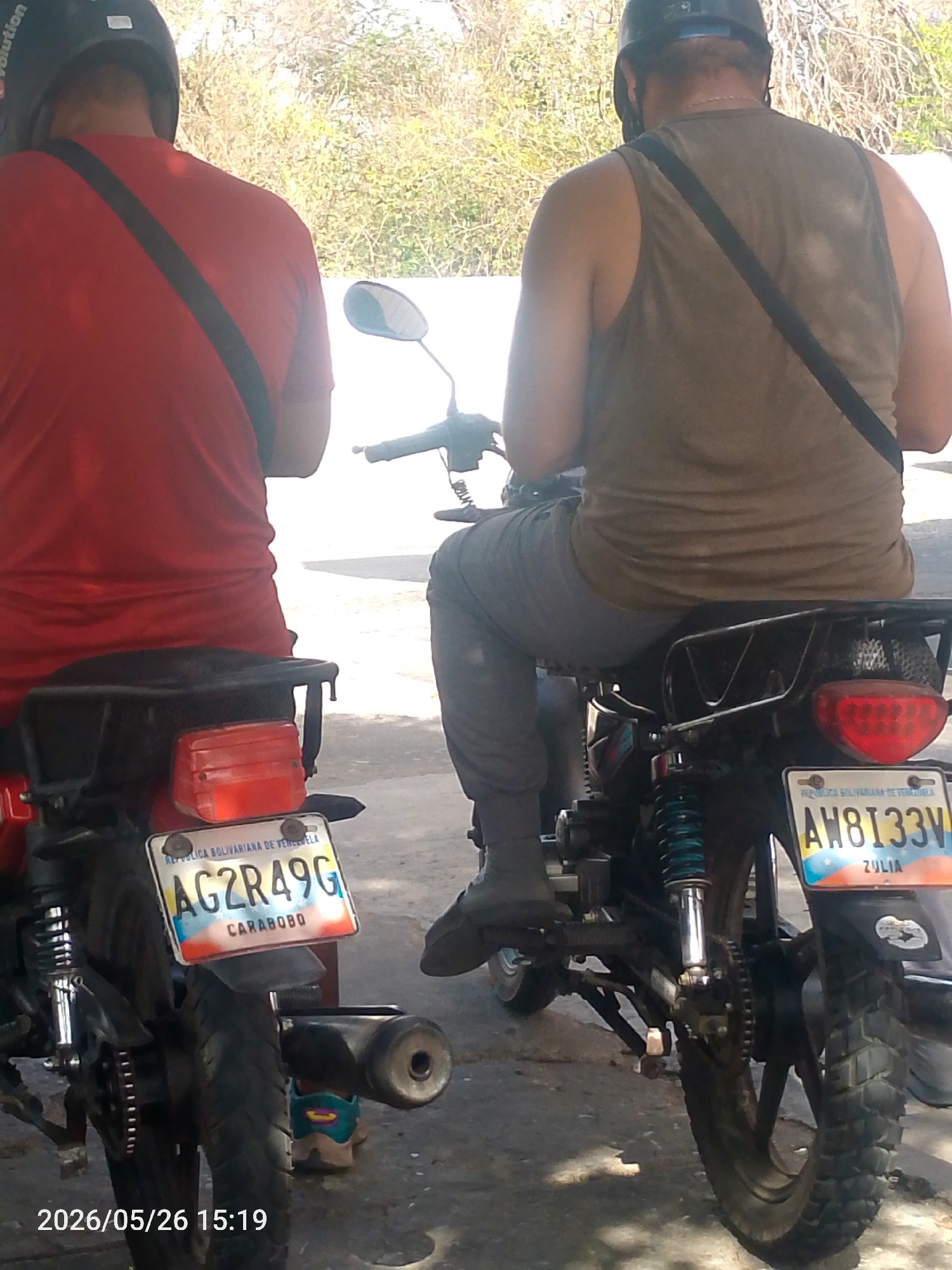 Two men are seated on motorcycles. The person on the left wears a red t-shirt and black helmet. The person on the right wears a tan tank top, gray pants, and a black helmet. Both have a black strap across their chests and backs.
The motorcycle on the left has a license plate reading "AG2R49G" and is from Carabobo, Venezuela. The motorcycle on the right has a license plate reading "AW8133V" and is from Zulia, Venezuela.
The image is taken during the day. The timestamp on the bottom reads "2026/05/26 15:19". The background consists of vegetation. The image was taken in La Tagua, Venezuela.