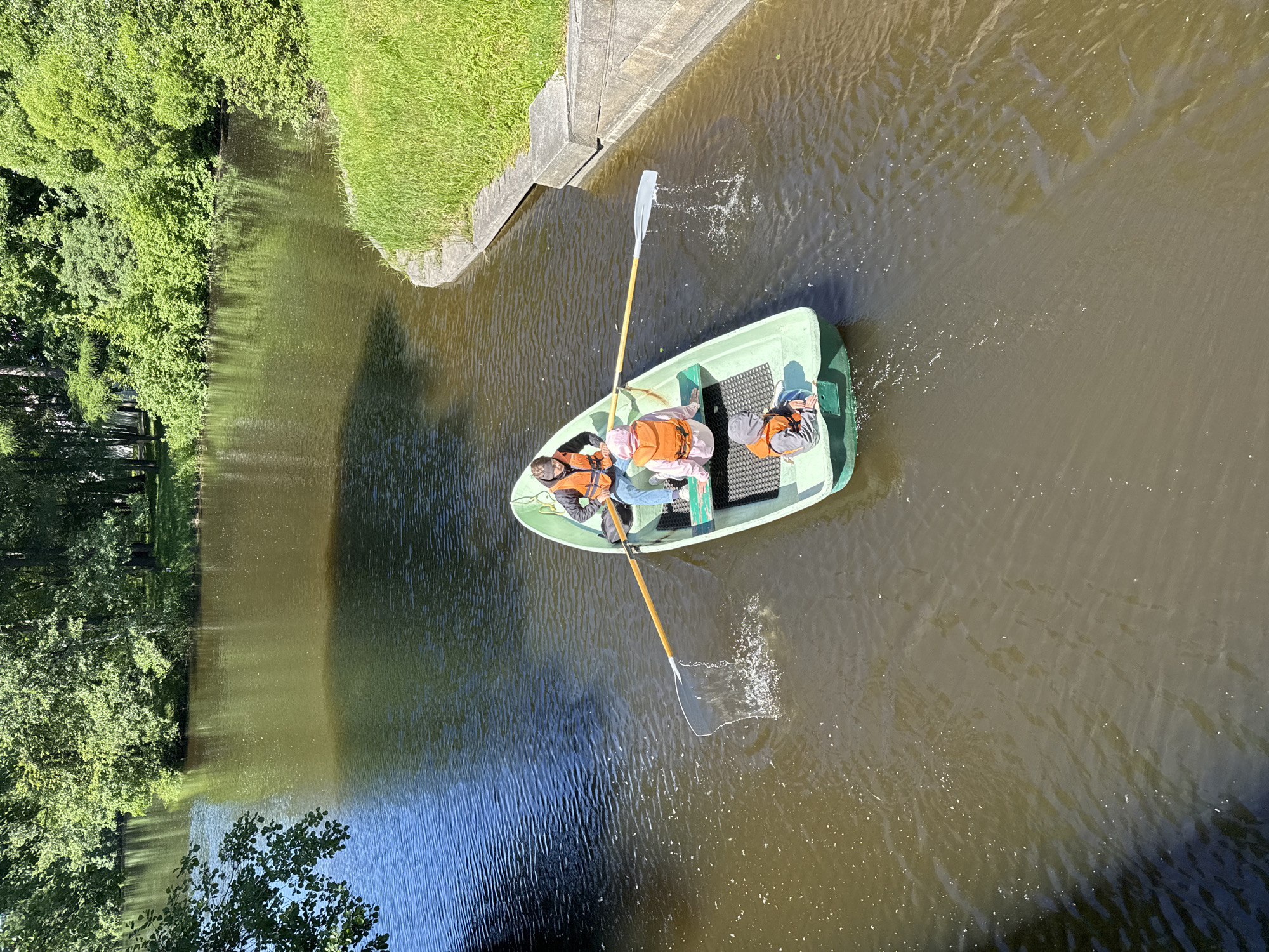 A light green rowboat with three individuals wearing orange life vests glides through a brown body of water. The person in the stern is rowing with two oars, one of which is partially submerged, creating a spray. The person in the middle is holding a fishing net that is also in the water. The third individual, a child, sits at the bow. The boat is in a canal or river lined with lush green trees and grass on both banks. A concrete embankment is visible on the right side of the frame. The water is calm with ripples reflecting the sunlight. The scene appears to be in a park or recreational area within Saint Petersburg, Russia, likely during daytime given the bright lighting and shadows.