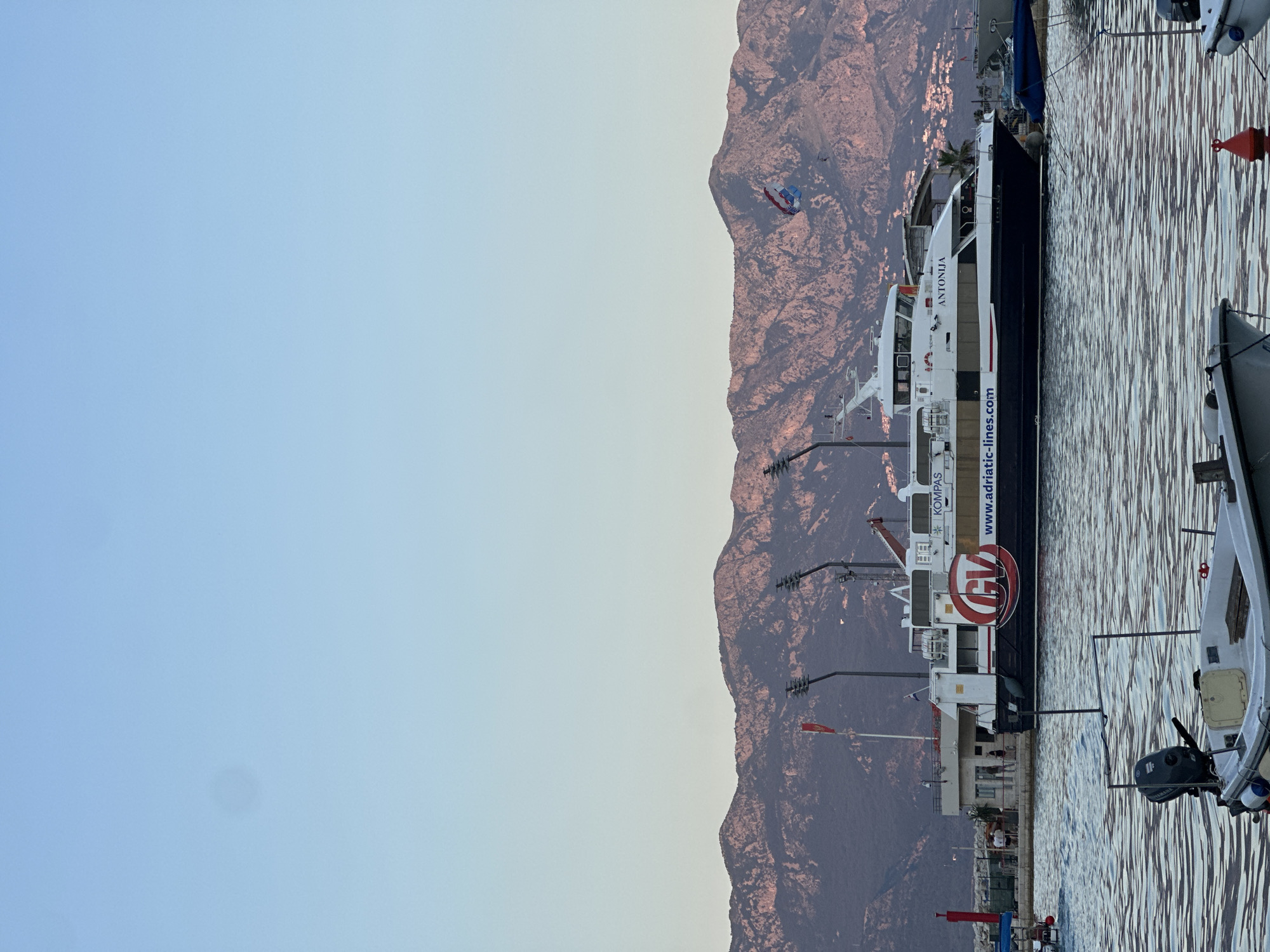 In Budva, Montenegro, a large ferry named "ANTONIJA" is docked at a port, with mountains in the background. The ferry has the "GV" logo and "www.adriatic-lines.com" written on its side. Several people are standing on the dock near the ferry. In the foreground, two small boats are visible on the water, one with an outboard motor. The sky is clear and blue, with a gradient towards a lighter blue near the horizon. The mountains are illuminated by the setting sun, casting a warm glow on their slopes.