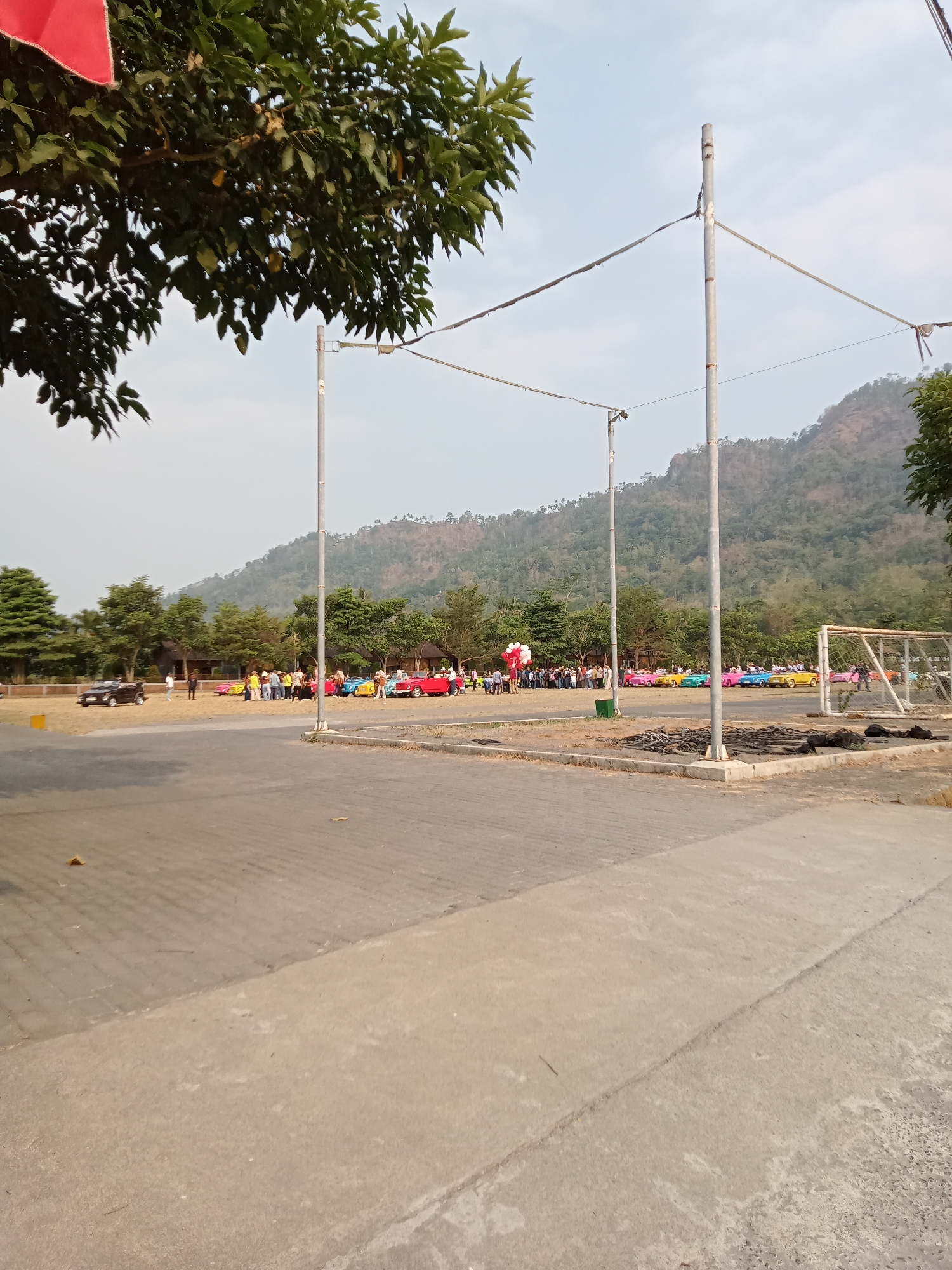 The image captures an outdoor scene in Majaksingi, Indonesia, featuring a gathering of people and a line of colorful classic cars. In the foreground, a paved area transitions to a darker, textured surface. In the middle ground, a group of individuals stands around a collection of brightly colored vehicles, including red, yellow, blue, and pink convertibles. Behind the cars, a cluster of trees forms a backdrop, with a few structures visible amongst them. Further in the distance, a mountainous landscape with sparse vegetation dominates the horizon under a hazy sky. The scene suggests a car show or community event.