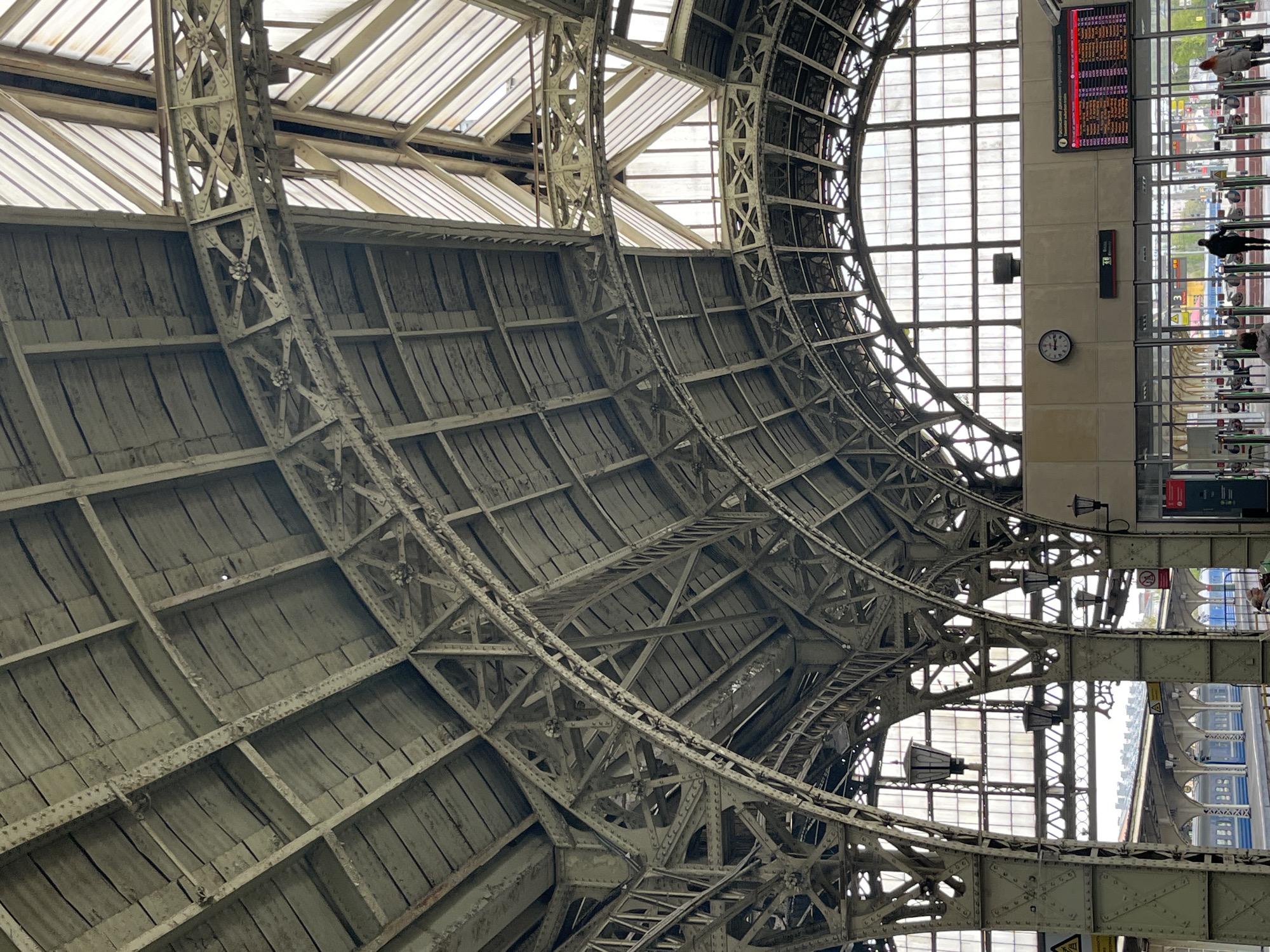 The image shows the interior roof structure of a train station. The roof is a curving structure of metal beams and wooden planks, with skylights near the top. To the right, there is a wall with a clock, a departure board, and people visible through a window. The city and specific location are unknown.