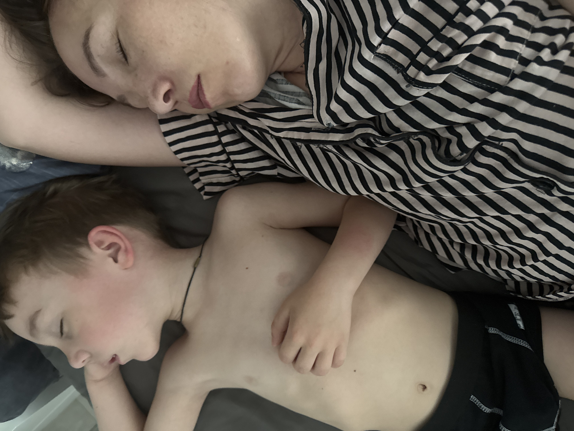 A woman and a child are sleeping in a bed. The woman is wearing a black and white striped top. The child is wearing black shorts and a necklace. They are lying on a gray sheet. The location is unknown; the city of Chaikovsky, Russia, cannot be confirmed from the image.