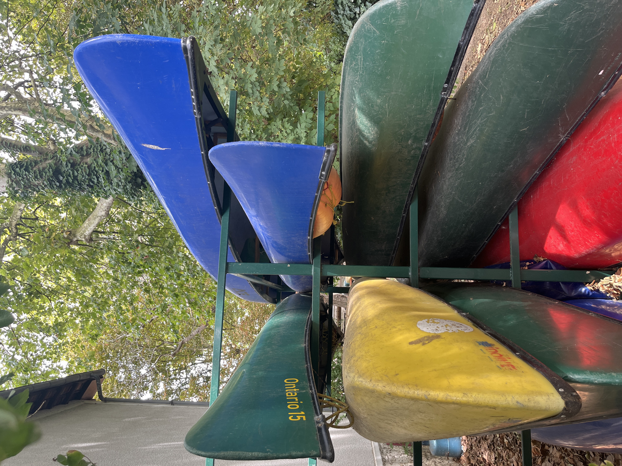 The image shows a rack of canoes stored outdoors. The canoes are of various colors including blue, green, yellow, and red. The rack is made of green metal and is positioned in front of a building with a grey exterior and a dark roof. Trees with green leaves are visible in the background, suggesting an outdoor setting. The scene appears to be in a recreational area, possibly a park or a watersports facility, located in Joinville-le-Pont, France. No people are visible in the image, and there are no ongoing activities depicted. The overall impression is one of organized storage of watercraft in a natural environment.