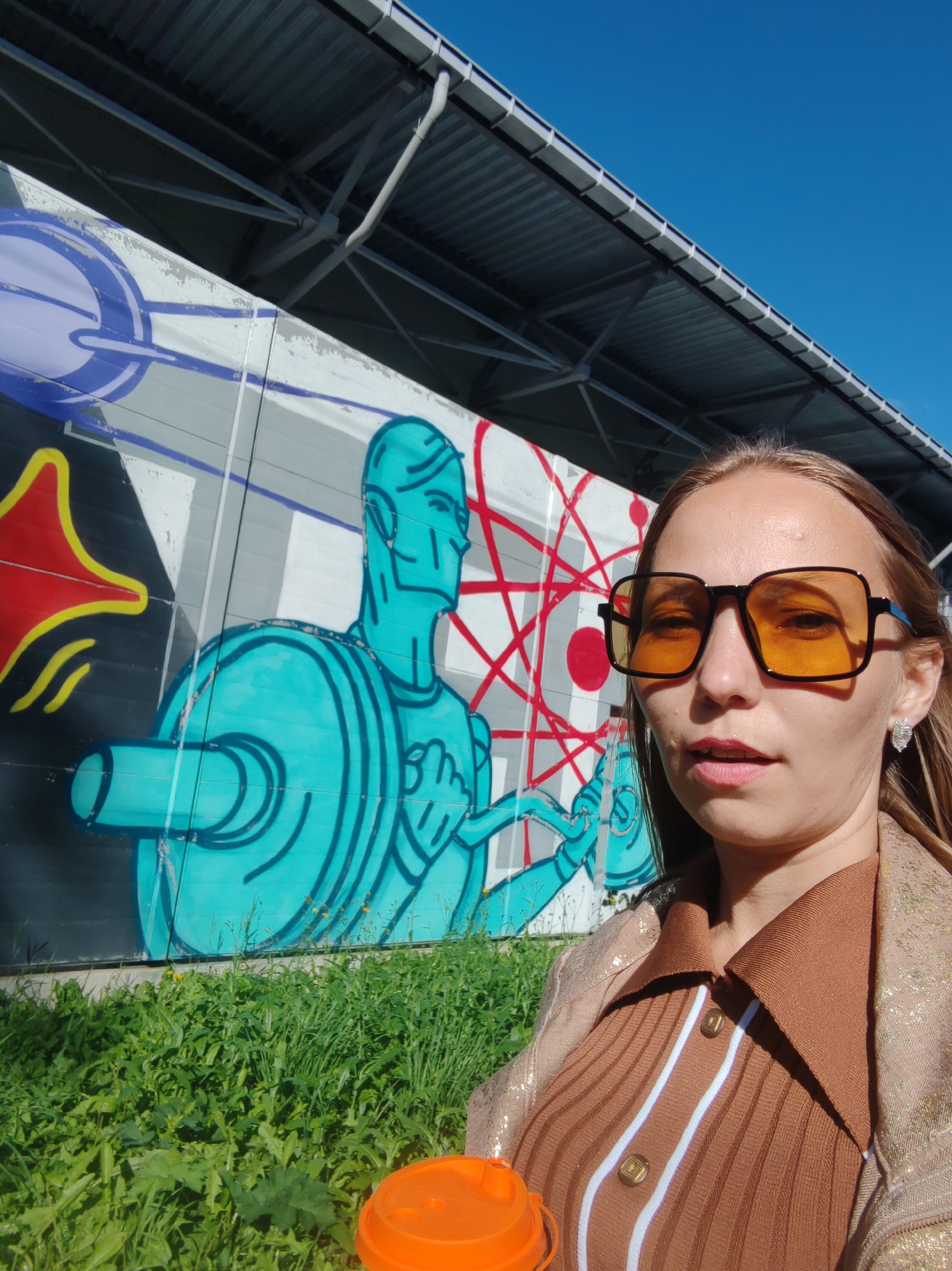 A young woman is taking a selfie outdoors in front of a mural. She has long, light brown hair and is wearing large, square, orange-tinted sunglasses, a brown ribbed shirt with white stripes and gold buttons, and a shimmering gold jacket. She is holding an orange lid in her right hand. The mural behind her depicts a stylized blue figure with a red and white geometric design. The background shows a blue sky, a gray roof structure, and green grass. The location is not identifiable as Syktyvkar, Russia.