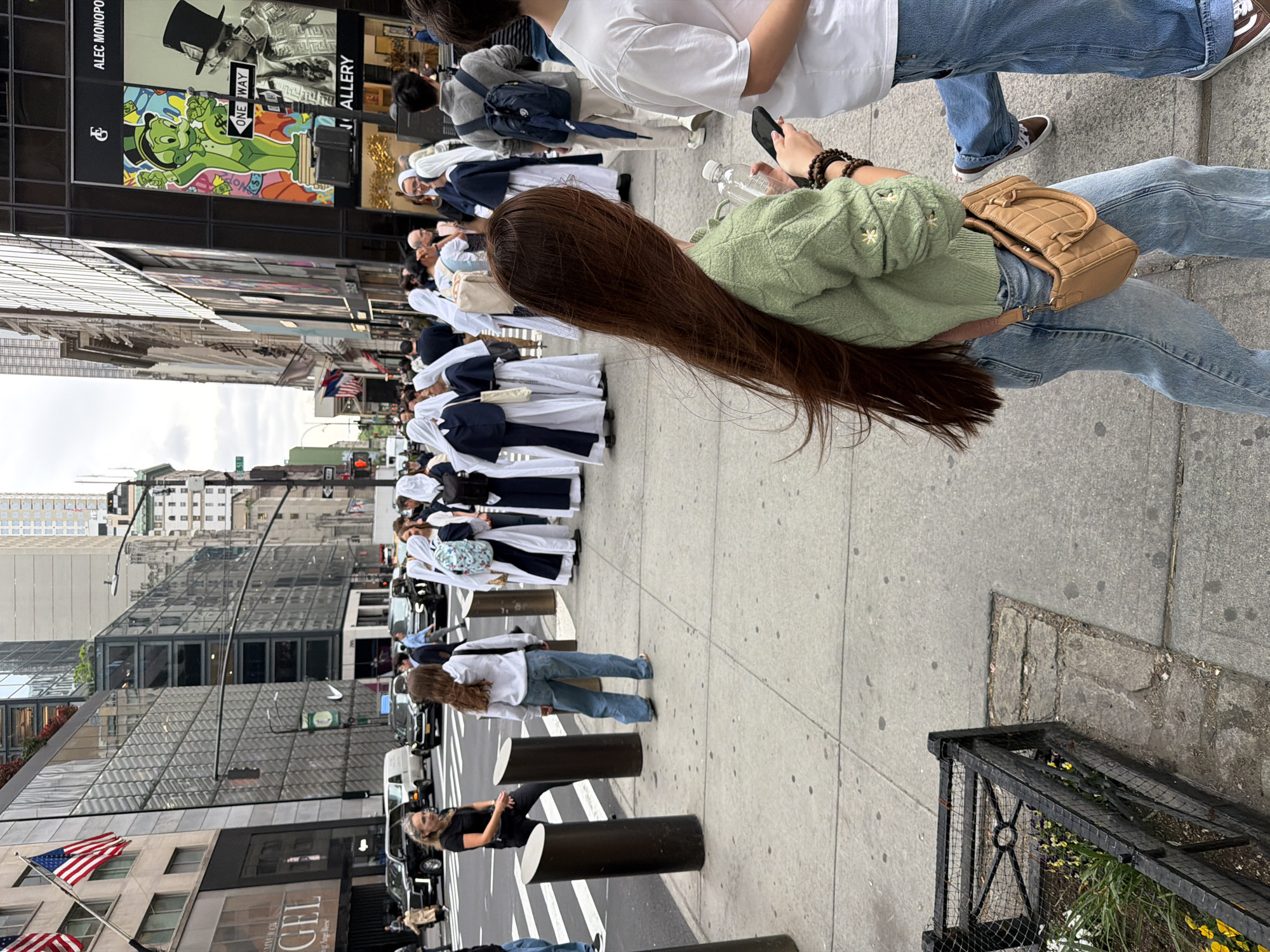 The image shows a street scene in New York City, United States of America. A group of women, dressed in white and navy blue religious habits, are walking along the sidewalk. A person with long brown hair, wearing a green sweater and blue jeans, is walking in the opposite direction, holding a cell phone. The sidewalk is paved with gray concrete and there are cylindrical black bollards along the street. The buildings in the background are tall and modern, with glass and steel facades. An American flag is visible on a pole in front of one of the buildings. The street appears to be busy with pedestrian traffic.