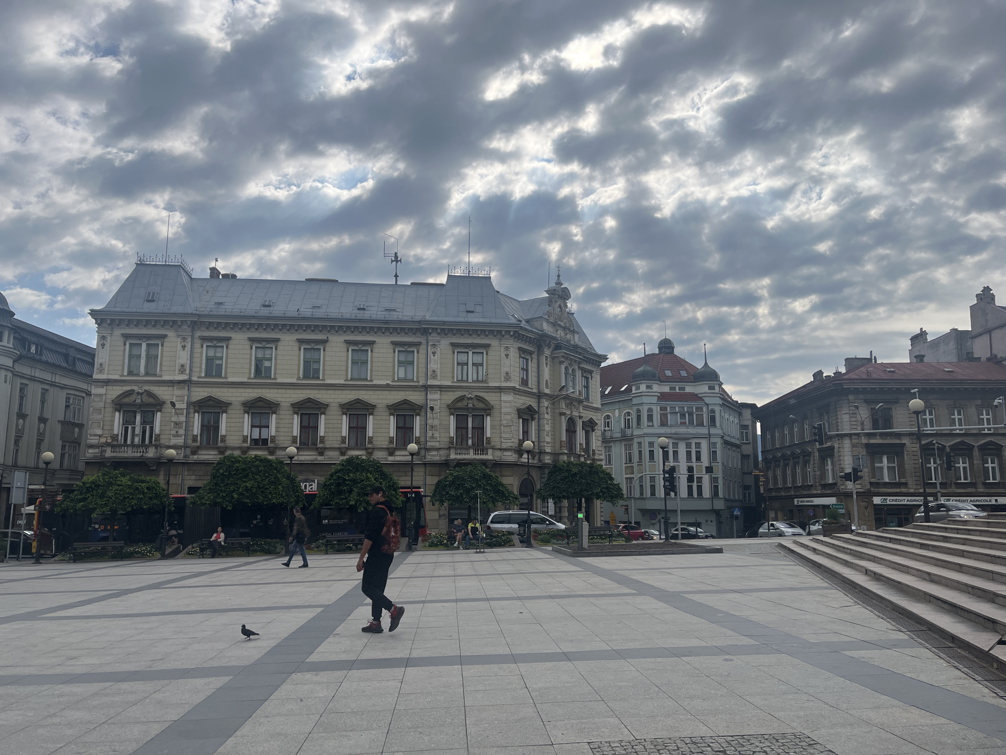 A wide-angle, eye-level shot captures a paved city square under a cloudy sky. The square features geometric grey and light grey tile patterns. Several people are present in the scene. In the foreground, a person walks from left to right, wearing dark clothing and a backpack. To the left, a lone pigeon stands on the tiles. Further back, near trees and benches, other individuals are seated or walking. On the left side of the square, an ornate building with multiple stories and many windows dominates the background, featuring a grey mansard roof with decorative elements. To the right of this building, a lighter colored, more modern-looking building with a teal roof is visible, followed by another older, darker building with multiple windows. To the far right, a set of wide concrete stairs ascends out of frame. Trees are planted in rows, forming a green border in the middle ground. The overall atmosphere is subdued due to the overcast conditions.