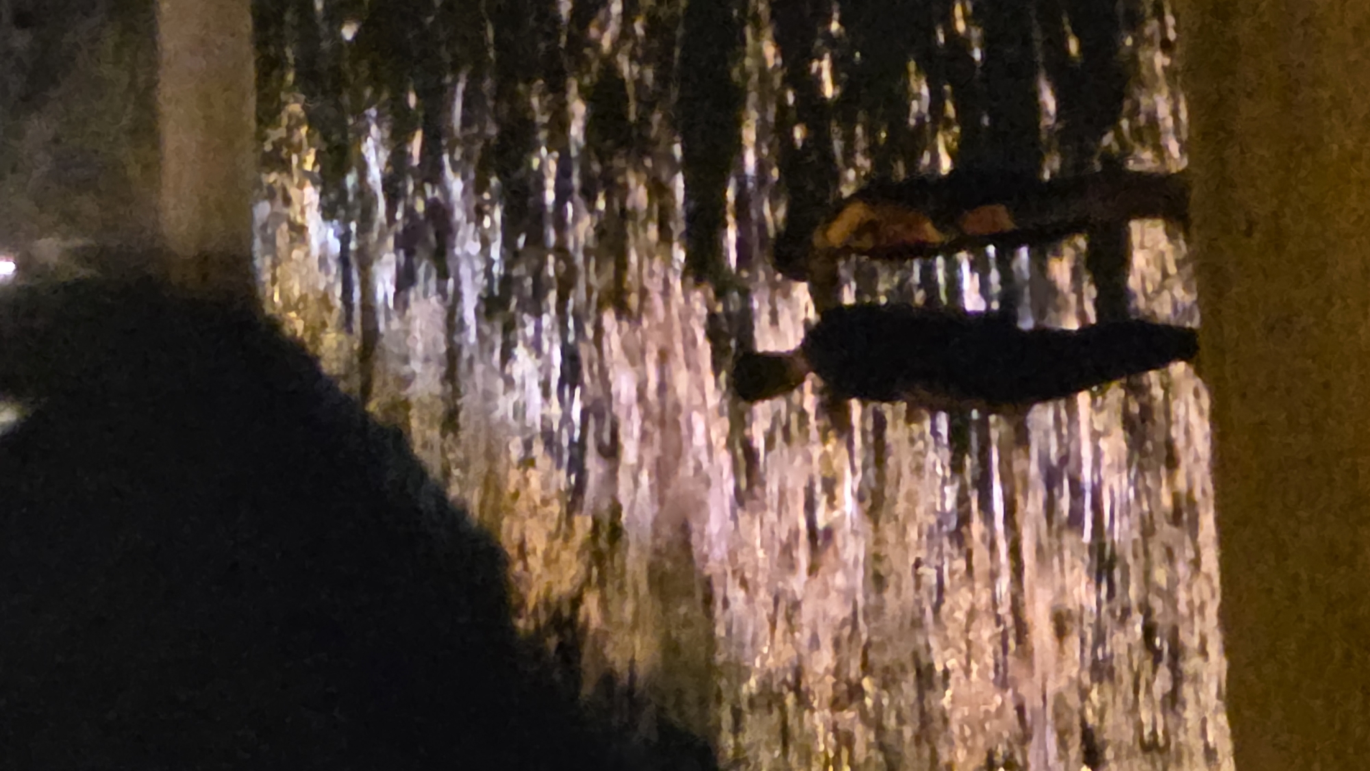 The image shows a blurry outdoor scene at night in Nha Trang, Vietnam. The main subject is a lighted decorative waterfall feature. Two dark figures are visible on a structure in front of the waterfall. To the left is a large, dark, indistinct shape. The overall lighting is dim and the image quality is poor, but it seems to be a public space.