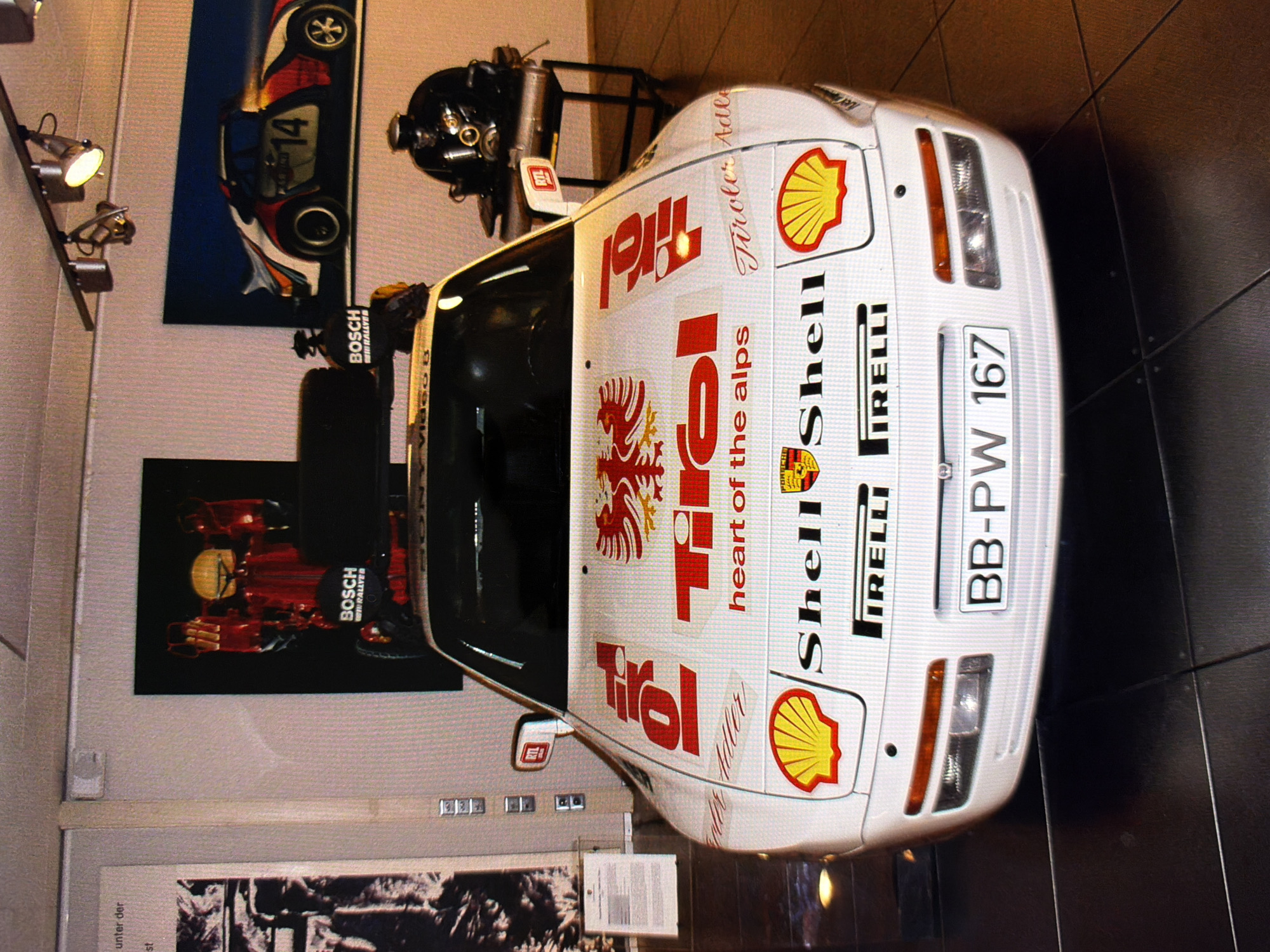 An eye-level, medium shot shows the front of a white Porsche 944 race car parked on a dark tiled floor indoors. The car has numerous sponsor decals, including red "Tirol" logos, an eagle crest, "Shell" logos with yellow shells, and "Pirelli" text. A license plate reads "BB-PW 167". To the left of the car, a poster with a red and yellow figure and "BOSCH" text is visible on a white wall. To the right, a framed picture of a white and red Porsche race car with the number "14" is displayed on a blue background. In the background, a black helmet sits on a stand with a silver exhaust pipe underneath. The lighting appears to be from spotlights. The scene is well-lit and the car is the primary subject.