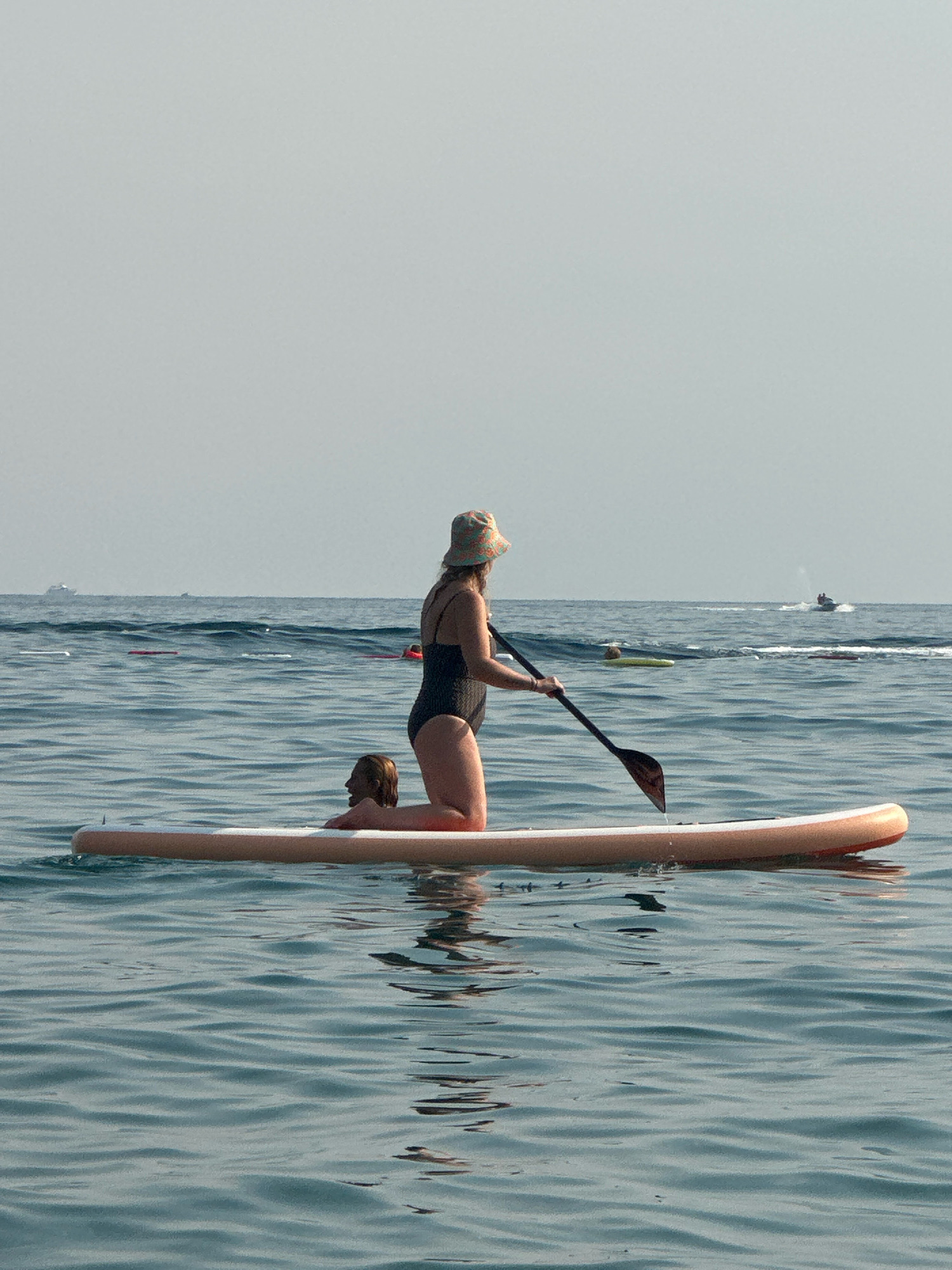 This media file captures a serene scene on the water in Boreti, Montenegro. Two people are on a stand-up paddleboard, with one woman kneeling and holding a paddle. She is wearing a dark swimsuit and a patterned hat. A second person, partially visible, is lying on the paddleboard behind her. The water is calm and reflects the clear, overcast sky, suggesting it is daytime with mild weather.
In the background, several other individuals are visible on the water, some on inflatable rafts and one person on a jet ski. A ship is also visible on the distant horizon, hinting at a popular recreational area. The overall mood is peaceful and relaxed, indicative of a leisurely day spent outdoors. No visible text is present in the image.