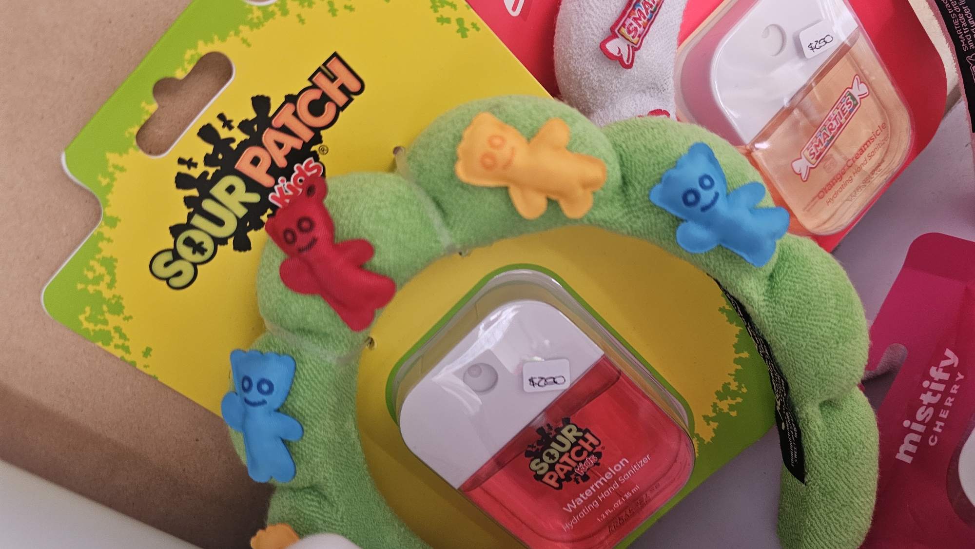 The image shows a variety of candy-themed personal care items. Included are: Sour Patch Kids hand sanitizer (watermelon scented), Smarties hand sanitizer (orange creamsicle scented), and a mistify cherry product. A green, plush head band with Sour Patch Kids characters attached is also present. The items are in a cluttered arrangement with a light brown background.