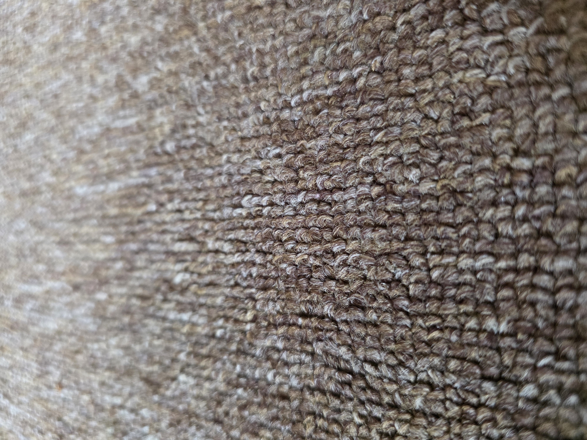 The media file is a close-up, high-angle shot of a textured surface. The surface appears to be made of tightly woven fibers, predominantly in shades of brown and white. The fibers are short and create a fuzzy, almost carpet-like texture. The lighting suggests a bright, indoor environment, with the light source casting subtle shadows that highlight the depth and texture of the material. The perspective is shallow, with the foreground in sharp focus and the background gradually blurring, creating a sense of depth. The composition is abstract, focusing entirely on the pattern and texture of the material rather than any specific objects or figures. There is no discernible activity or event taking place; the image is a static, textural study. No text is visible. Given the provided location context, the material could be a rug or upholstery found in a home or public space in Kyiv, Ukraine.