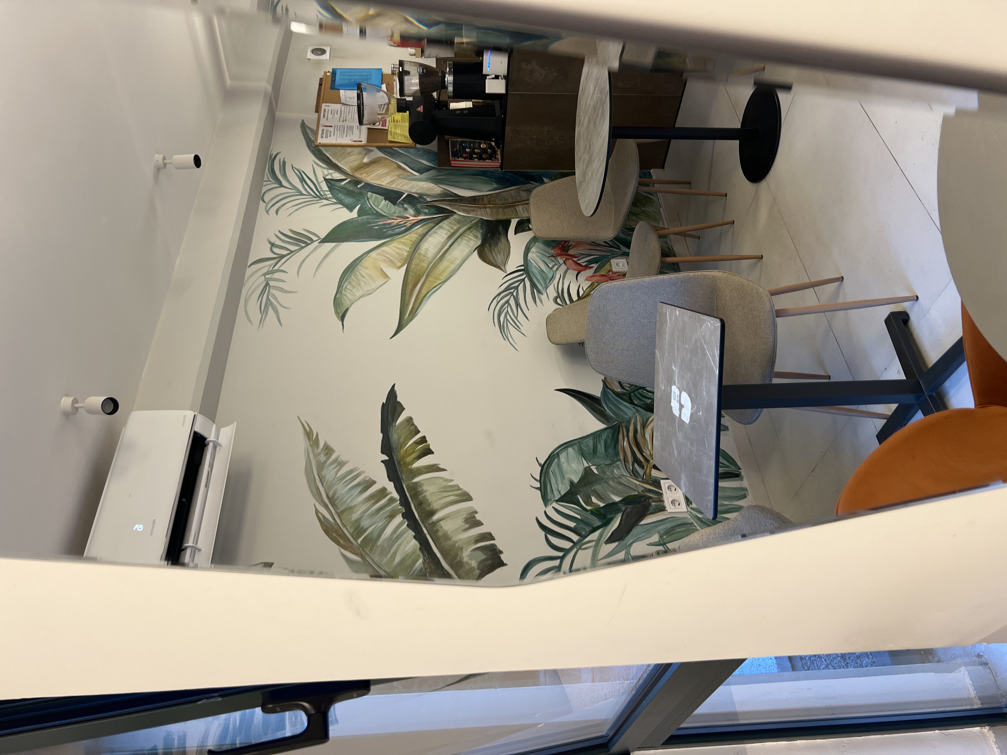 This is a photograph of the interior of a modern cafe, likely located in Budva, Montenegro, as indicated by the context. The scene is characterized by a clean, minimalist aesthetic with a tropical-themed mural on the wall.
In the foreground, a white pillar partially obscures the view, reflecting the light from the exterior. To the left, a large window offers a glimpse of the outside. The main area of the cafe features several small, dark marble-topped tables with black bases. Around these tables are various chairs, including upholstered grey chairs with wooden legs and a bright orange upholstered chair in the lower right corner.
The wall behind the tables is adorned with a vibrant mural depicting large, lush green tropical leaves and some red flowers, creating a striking contrast with the neutral tones of the furniture and the light-colored tiled floor.
In the background, a white air conditioning unit is mounted high on the wall. Nearby, a white spotlight fixture is attached to the ceiling. Further back, on a dark wooden counter, a professional coffee grinder and a coffee machine are visible, suggesting the cafe's function. A corkboard with various notices is also mounted on the wall. Electrical outlets are visible on the lower part of the muraled wall. The overall impression is of a contemporary, stylish cafe designed for relaxation and enjoyment.