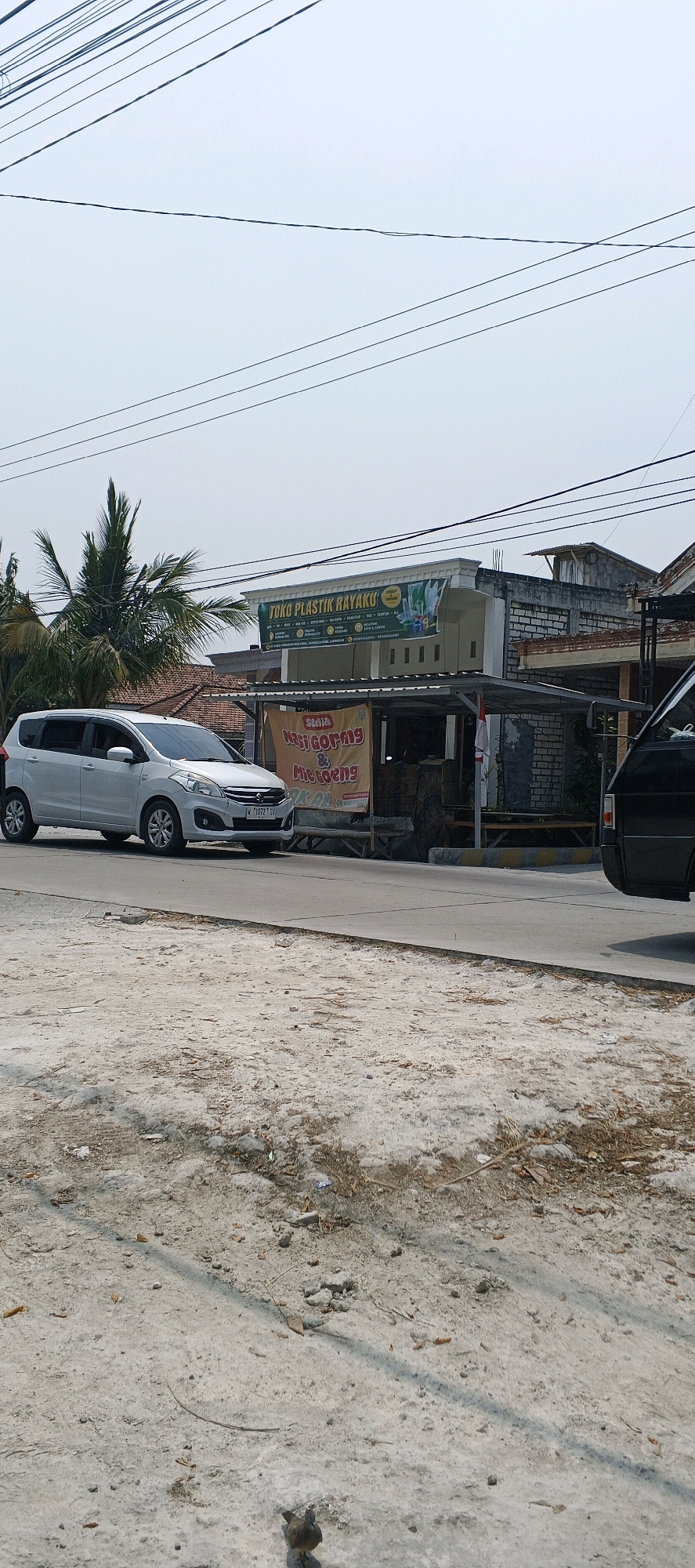 In Sonoadi, Indonesia, a silver minivan is captured in motion on a paved road. The vehicle, a Suzuki Ertiga, exhibits a license plate with the identifier "W 1072 SV". Adjacent to the road, a roadside stall displays a banner advertising "Nasi Goreng & Mie Goreng" (Fried Rice & Fried Noodles). Behind the stall, a building displays a banner for "TOKO PLASTIK RAYAKU," indicating a plastic goods store. Palm trees are visible in the background, alongside a clear, overcast sky. The ground in the foreground is a sandy, dirt surface. A small bird is present on the ground in the lower right portion of the image. Power lines are strung across the upper portion of the frame.