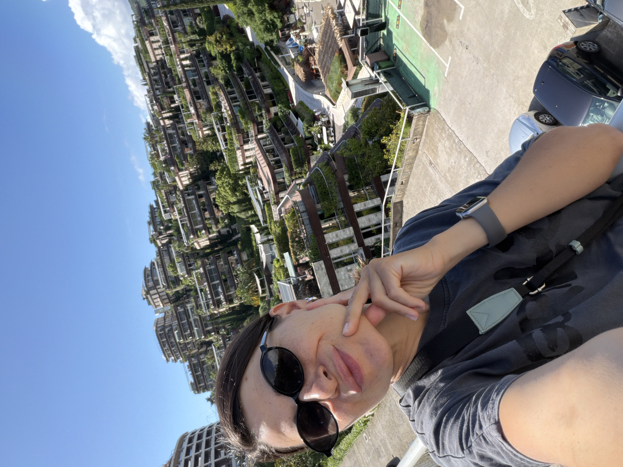 The image is a selfie captured by a person from a high vantage point overlooking a unique architectural complex, likely in Boreti, Montenegro, under a clear blue sky.
The main subject is an individual, likely an adult, seen from the chest up, with dark hair and wearing black sunglasses. They are dressed in a dark t-shirt with a subtle camouflage pattern and have a gray smartwatch on their left wrist. A dark strap, possibly from a bag, crosses their chest, adorned with a small, light green tag. The person has a playful expression, with a slight pout and one hand raised, fingers touching their cheek.
In the background, a striking series of terraced buildings are visible, characterized by extensive integration of lush green vegetation, resembling vertical gardens. These multi-story structures appear to follow the contours of a hill or slope. The sky above is a vibrant blue with a few scattered white clouds, indicating a bright and sunny daytime setting.
To the immediate right of the person, a concrete area with painted white lines and green sections, possibly a parking lot or a sports court, is discernible. Two cars, one dark and one lighter, are partially visible in this area. A railing separates the person from the ground below, suggesting they are on a balcony or rooftop. There is no visible text in the image.