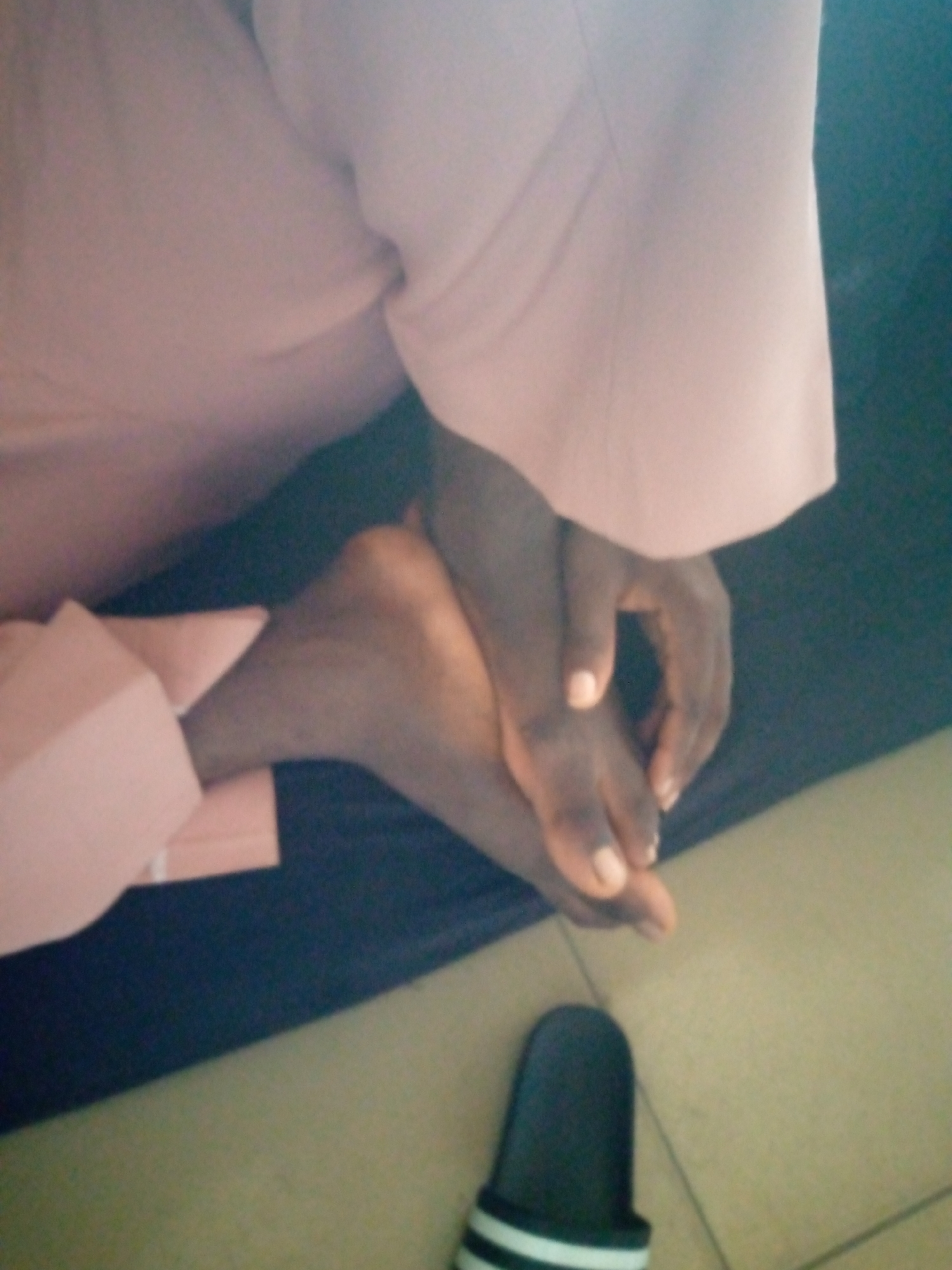 The image shows a close-up view of a person's feet and hands. The person appears to be wearing a light pink garment, possibly a robe or dress, and is resting their feet on a dark surface. One foot is visible, with toes extended. The hands are clasped together, resting near the feet. The background is dark, with a tiled floor visible in the lower right corner. A black slipper with white stripes is also present on the floor. The image does not provide any specific details that would allow for an accurate determination of its location within Jalingo, Nigeria.